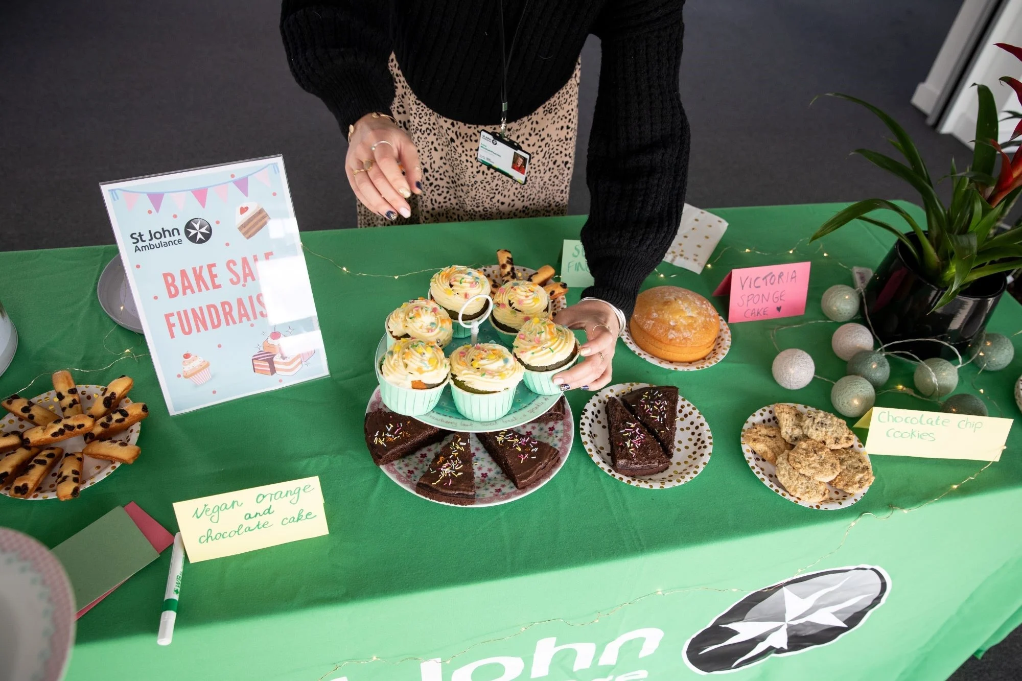 A table with a green St John Ambulance table cloth on it. It has a mix of difference cakes, biscuits, and cupcakes laid out on plates. It's also decorated with fairy lights and a plant, and has a sign saying 'Bake sale fundraising'. A person is stood behind the table picking up a cupcake.