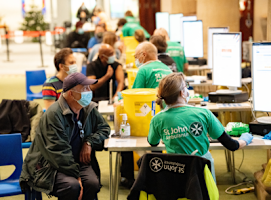 A photograph of SJA volunteers and patients at a vaccination centre. A line of tables stretch into the background, with a volunteer and patient sat at each one. Everyone is wearing face masks.