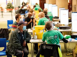 A photograph of SJA volunteers and patients at a vaccination centre. A line of tables stretch into the background, with a volunteer and patient sat at each one. Everyone is wearing face masks.