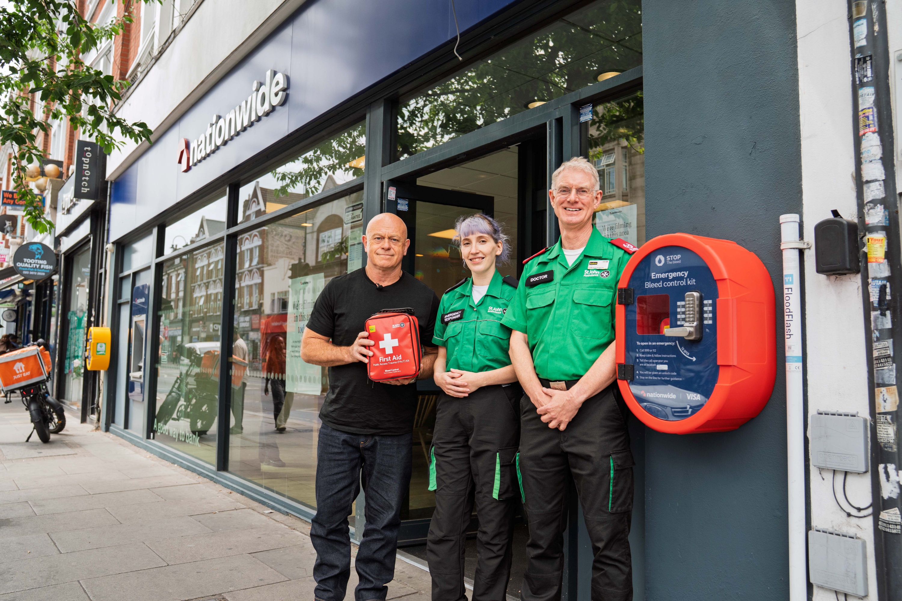 Ross Kemp and two St John Ambulance volunteers stood in front of a Nationwide branch. Toss Kemp is holding a defibrillator (AED).