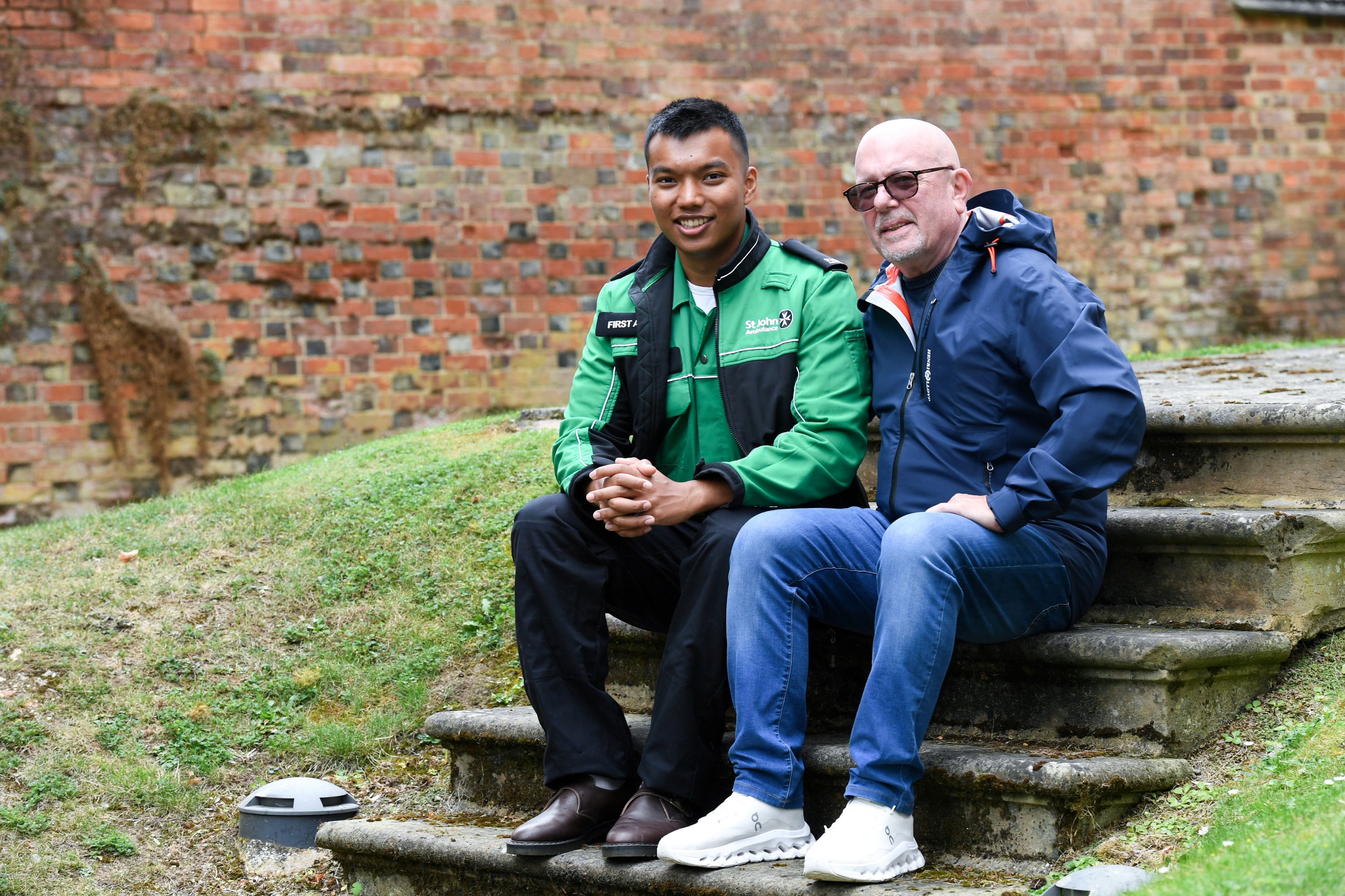 A photograph of Prince Tandukar and Paul Archre sitting outside, next to each other on some stone steps. Prince is wearing his St John Ambulance uniform.