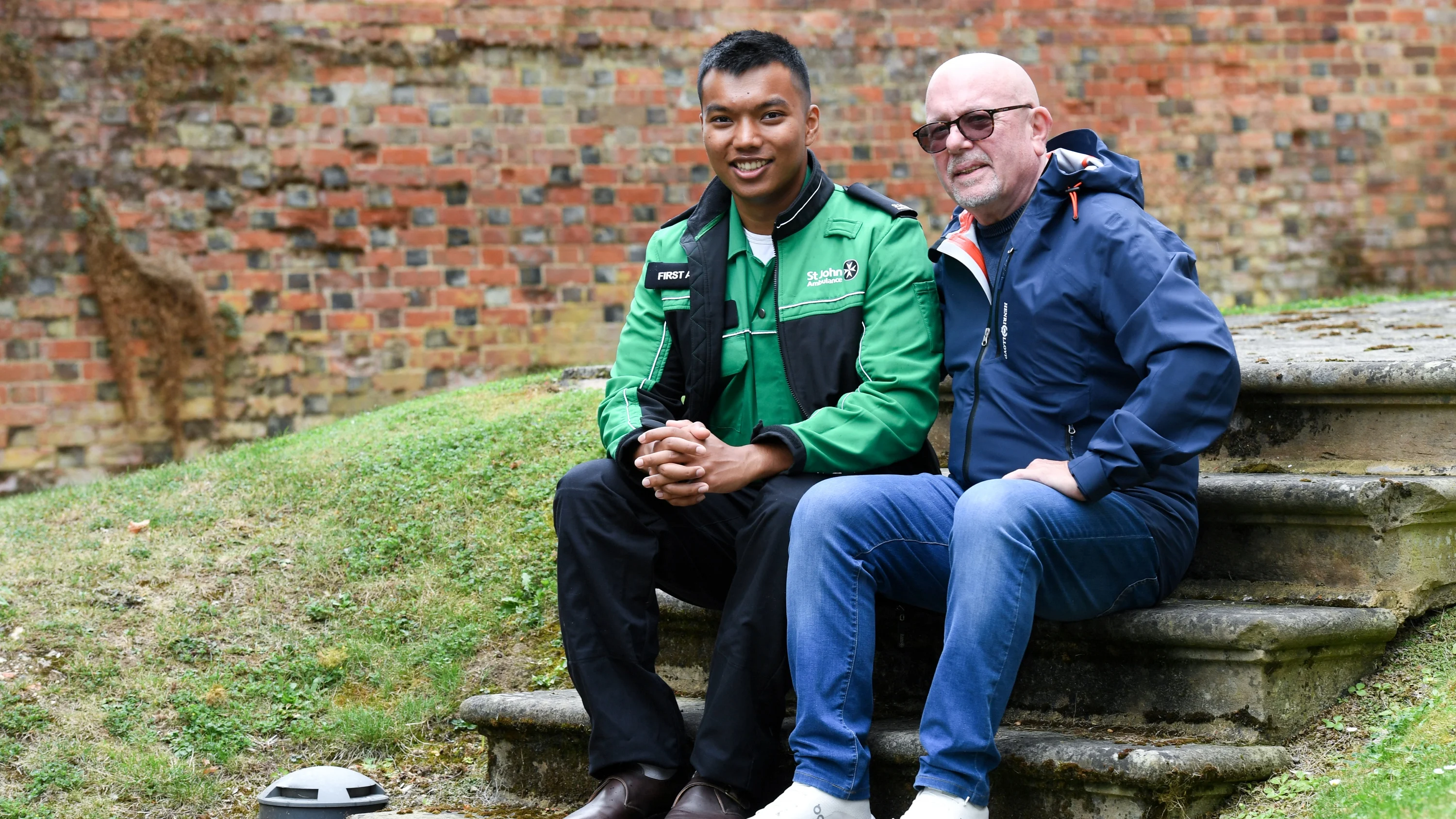 A photograph of Prince Tandukar and Paul Archre sitting outside, next to each other on some stone steps. Prince is wearing his St John Ambulance uniform.