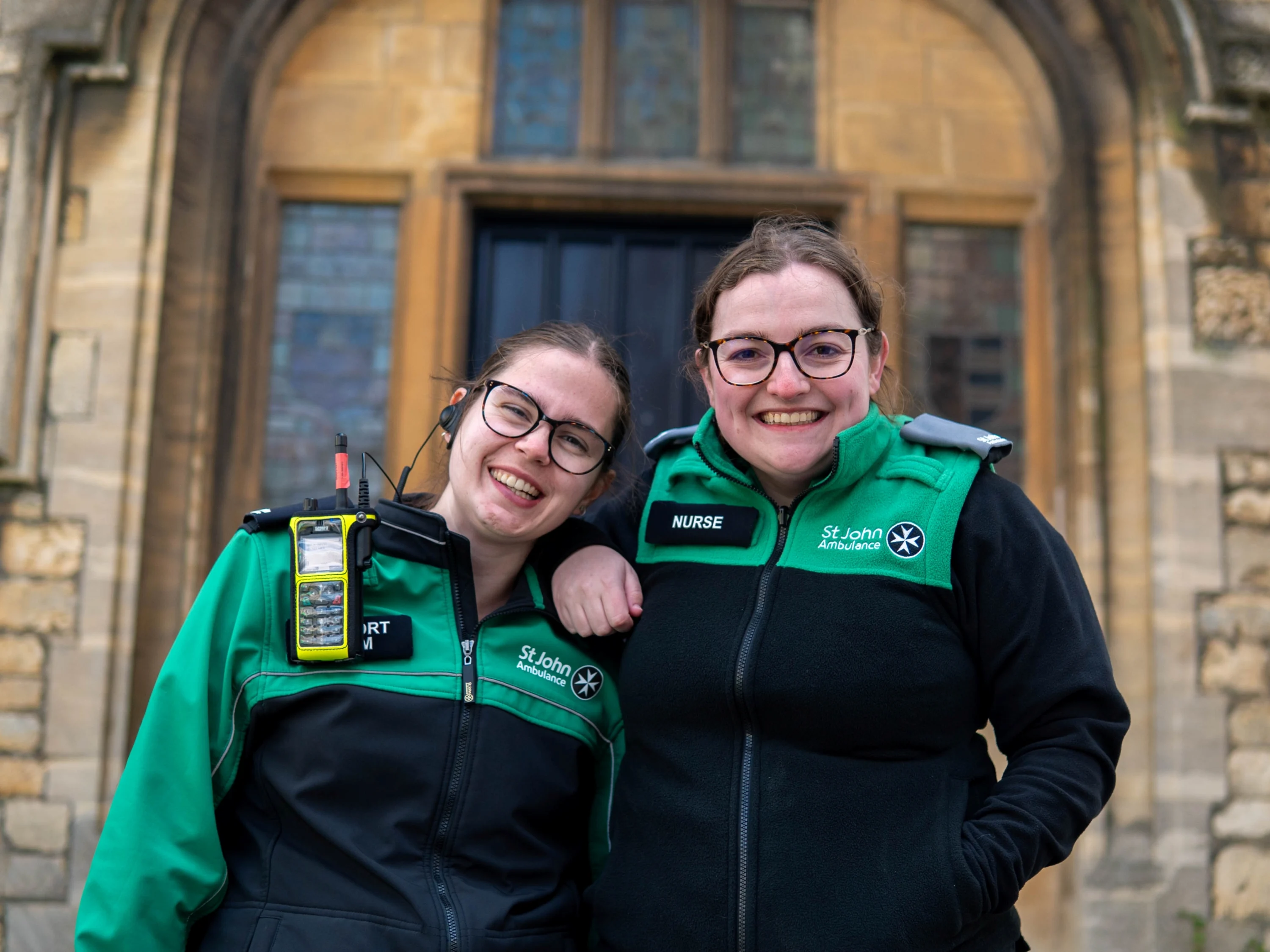 Two smiling St John Ambulance volunteers stood in front of an old building. One has a badge which says 'Nurse' and is resting their arm on the other's shoulder.