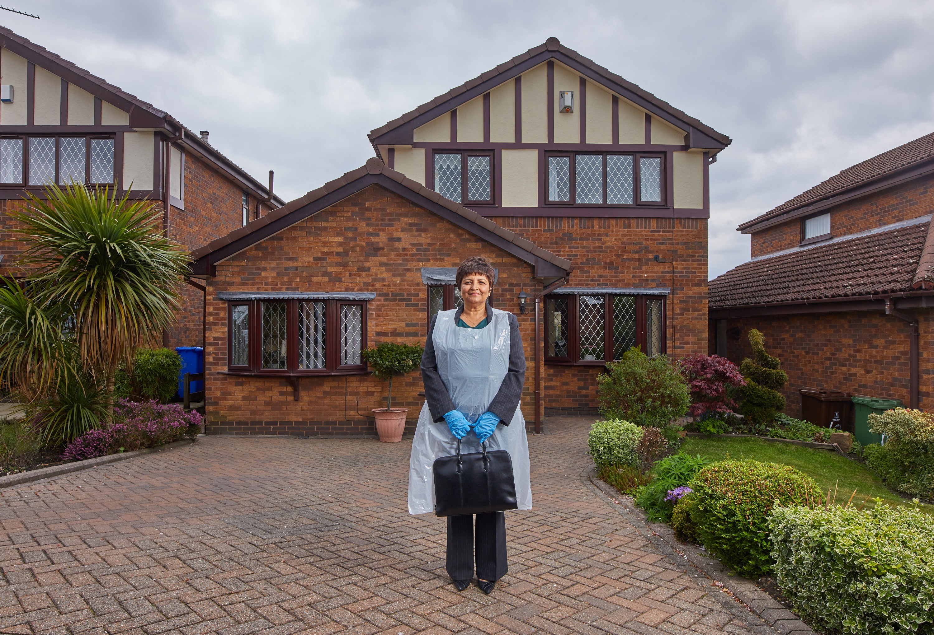 A photograph of Muni standing in her front garden, wearing PPE and holding a briefcase.