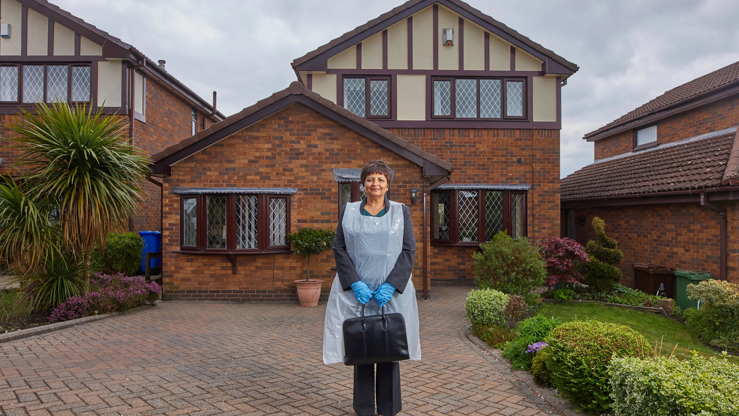 A photograph of Muni standing in her front garden, wearing PPE and holding a briefcase.
