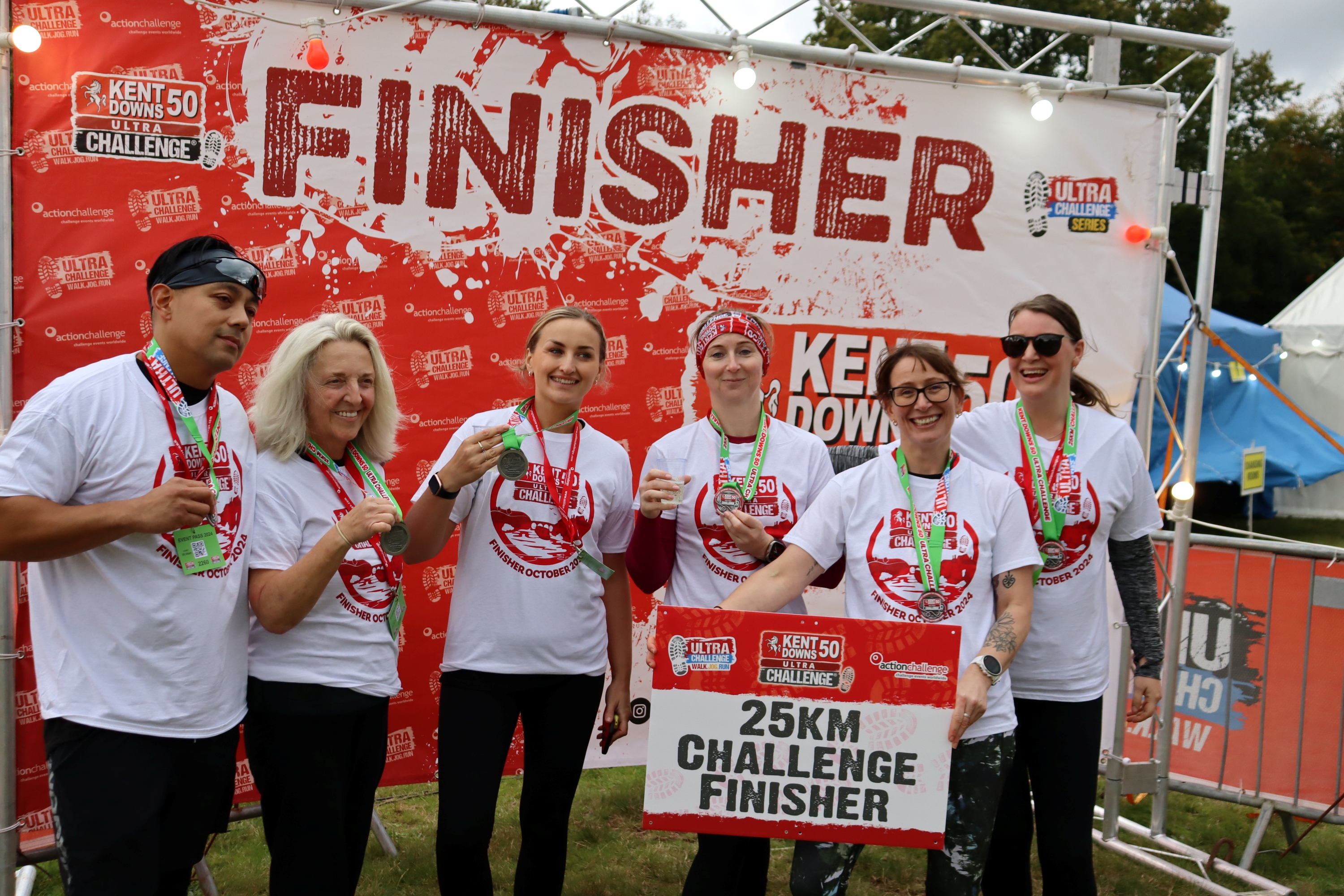 A small group of people that have completed the Kent Downs 50 Ultra Challenge, celebrating in front of a banner that has 'Finisher' on it.