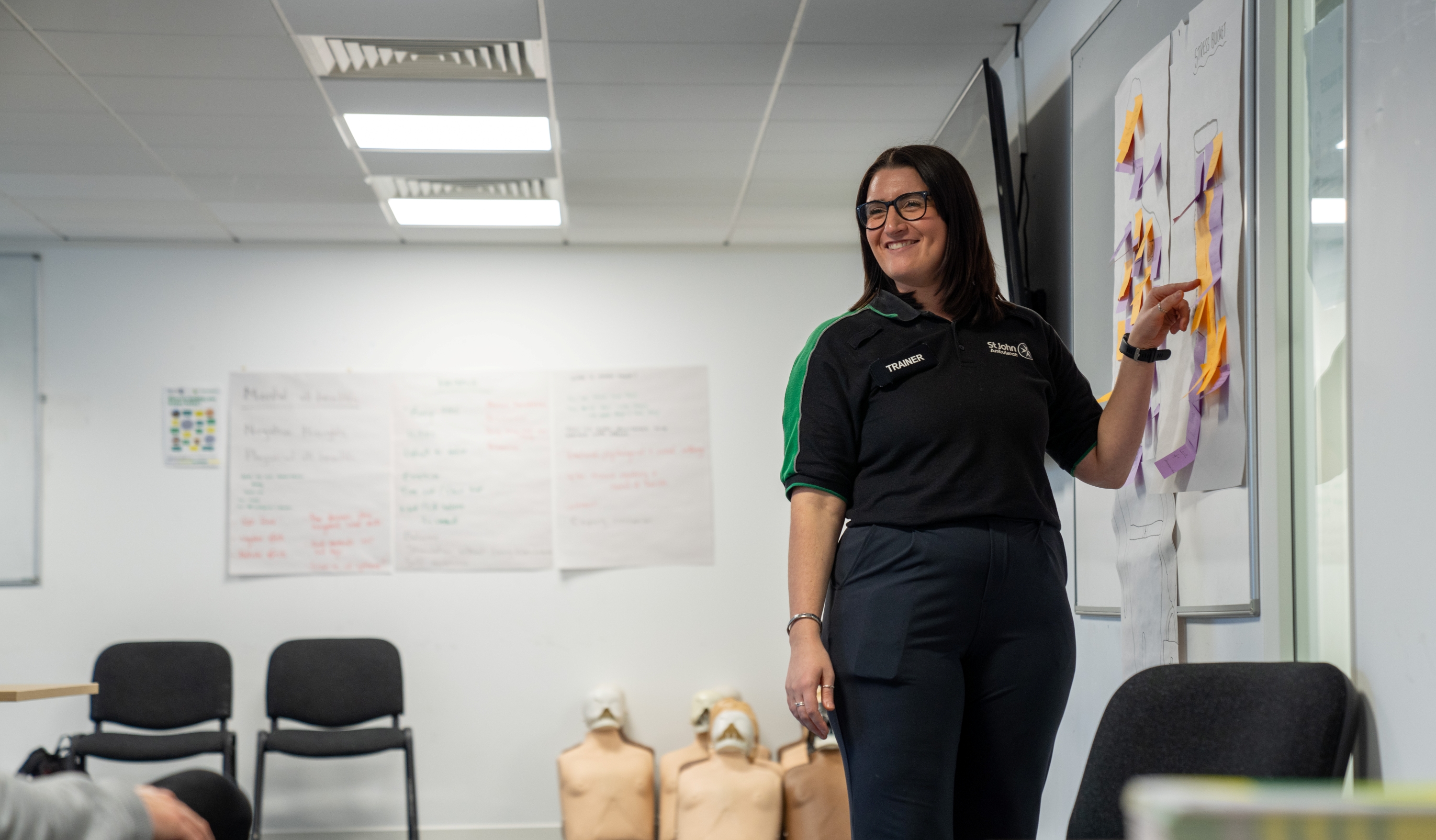 A smiling First Aid Trainer standing in a classroom pointing at some post its on a whiteboard. Behind them are some chairs and CPR mannequins as well as more large posters with writing on them stuck to the wall. 