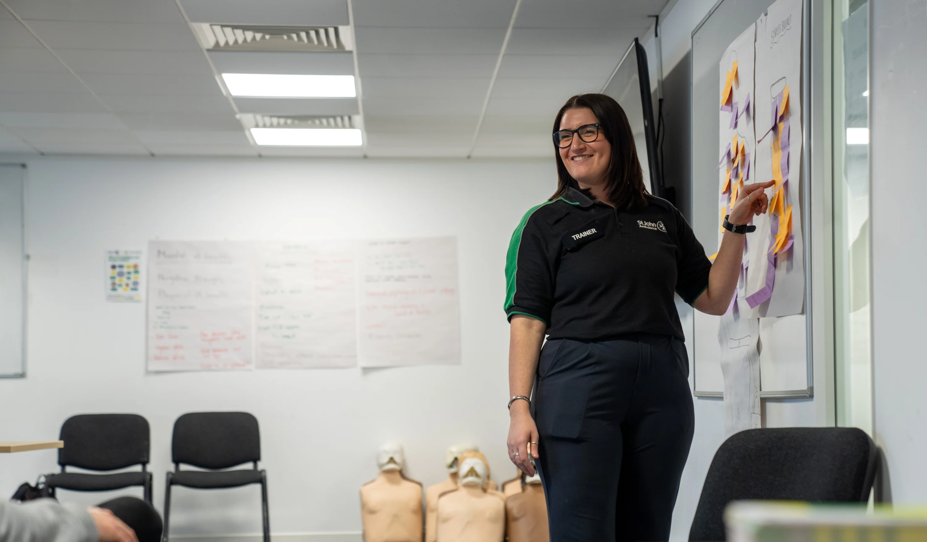 A smiling First Aid Trainer standing in a classroom pointing at some post its on a whiteboard. Behind them are some chairs and CPR mannequins as well as more large posters with writing on them stuck to the wall.