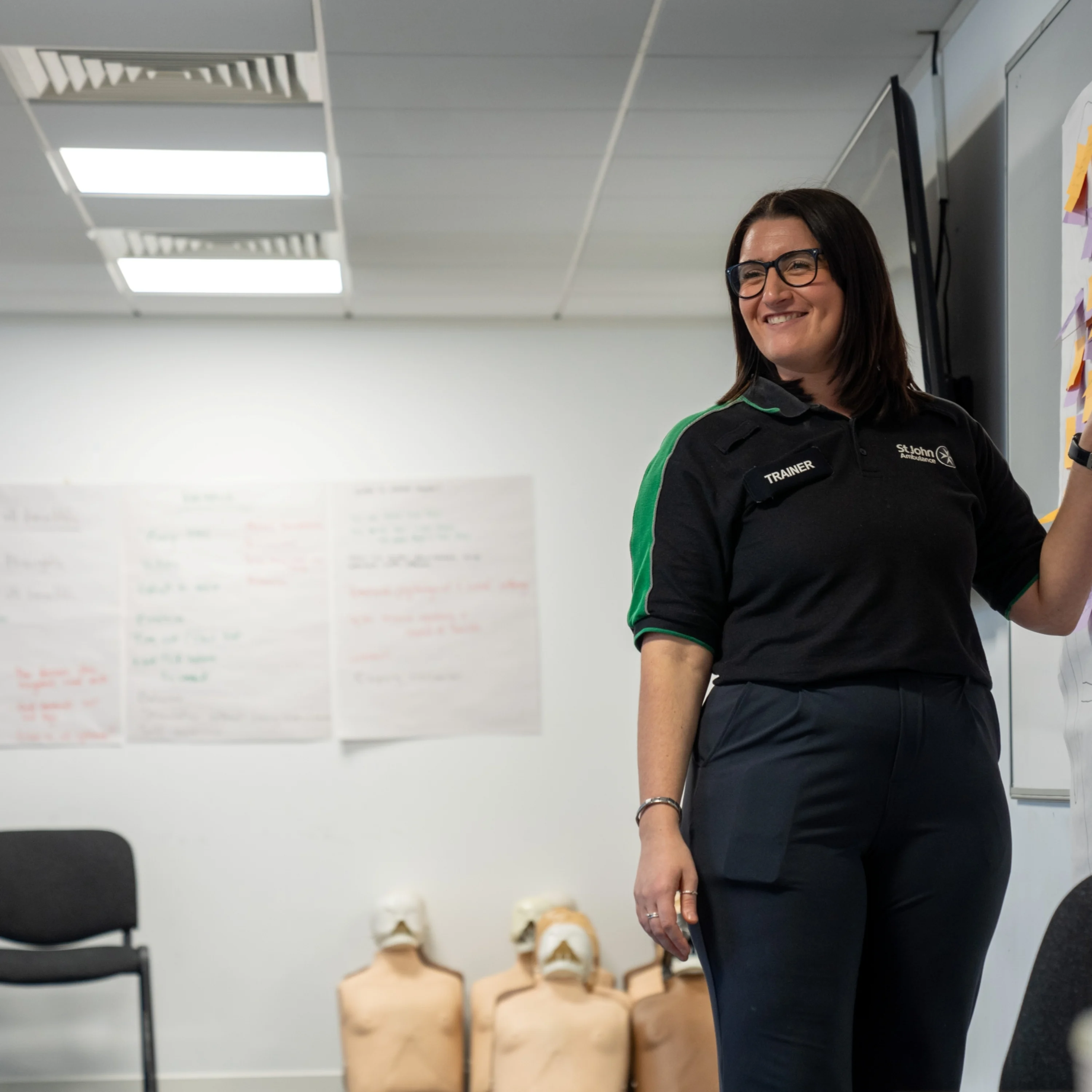 A smiling First Aid Trainer standing in a classroom pointing at some post its on a whiteboard. Behind them are some chairs and CPR mannequins as well as more large posters with writing on them stuck to the wall.