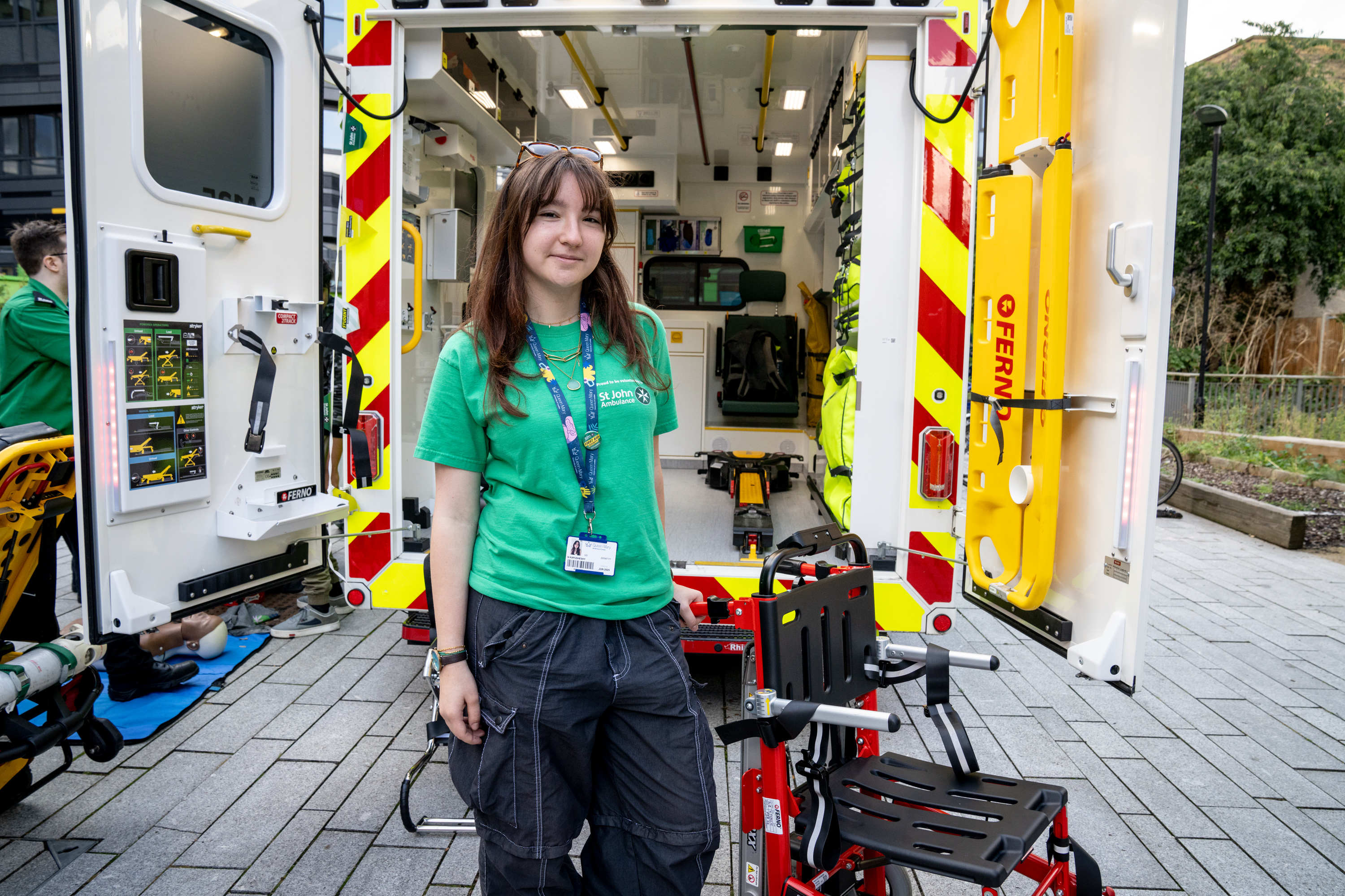 A photograph of a student volunteer standing in front of the back of an ambulance, with the doors open so you can see inside.