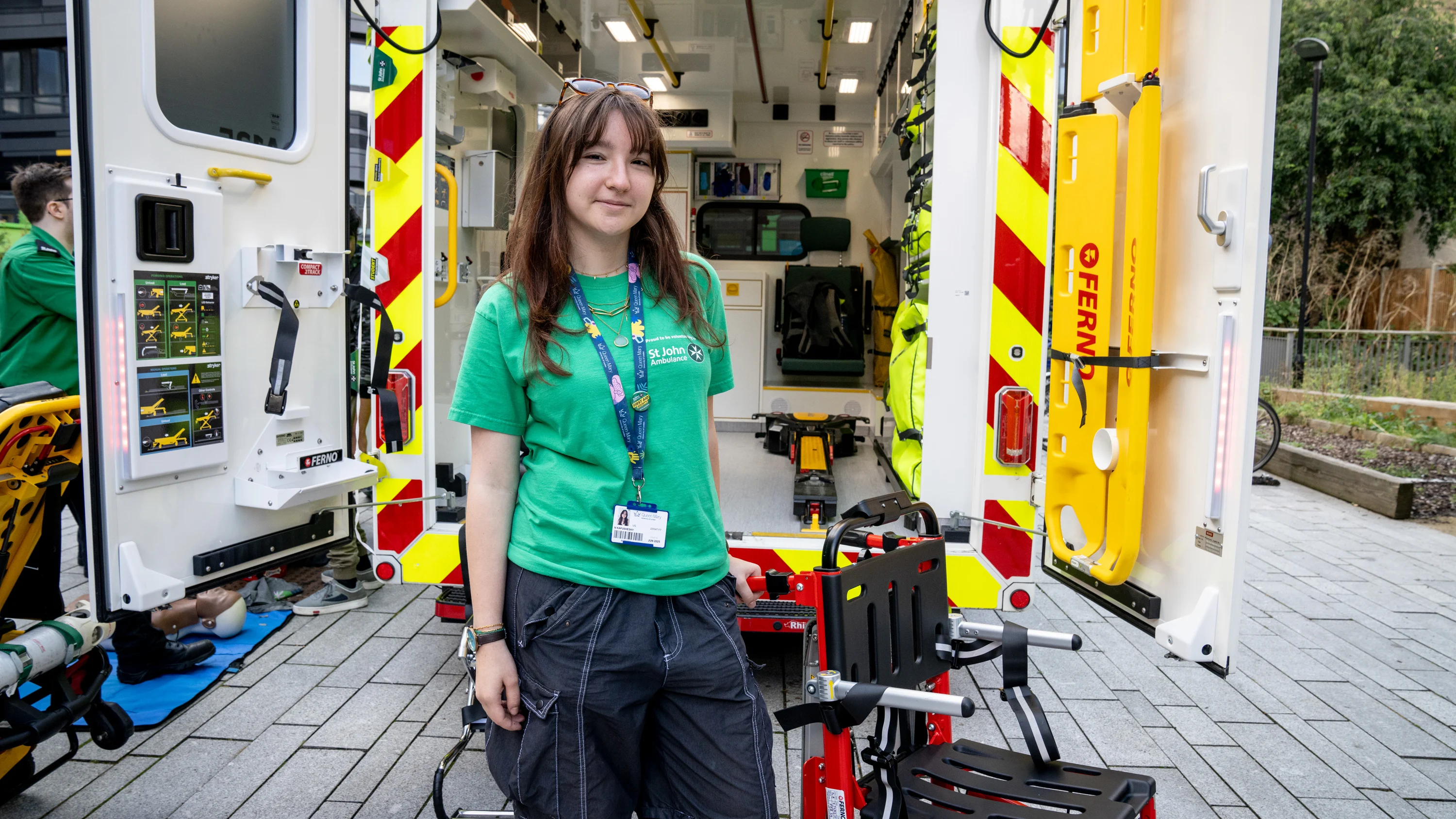 A photograph of a student volunteer standing in front of the back of an ambulance, with the doors open so you can see inside.