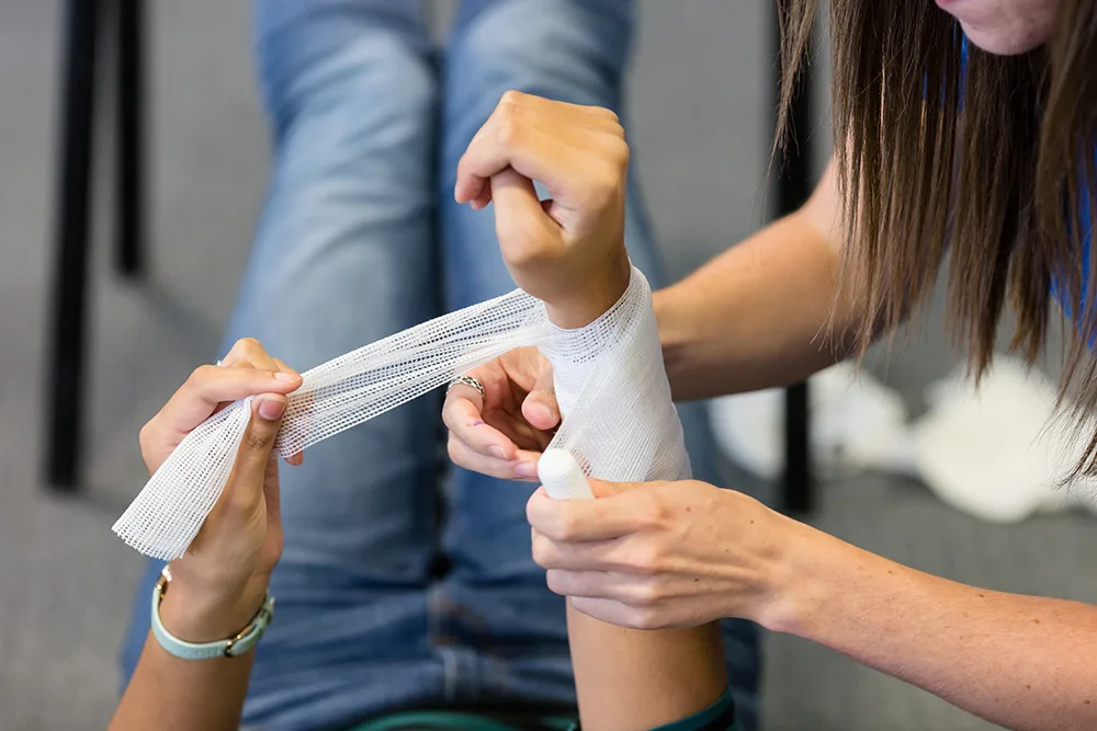 Someone wrapping a bandage around a person's wrist who is also lying on the floor with their legs up.