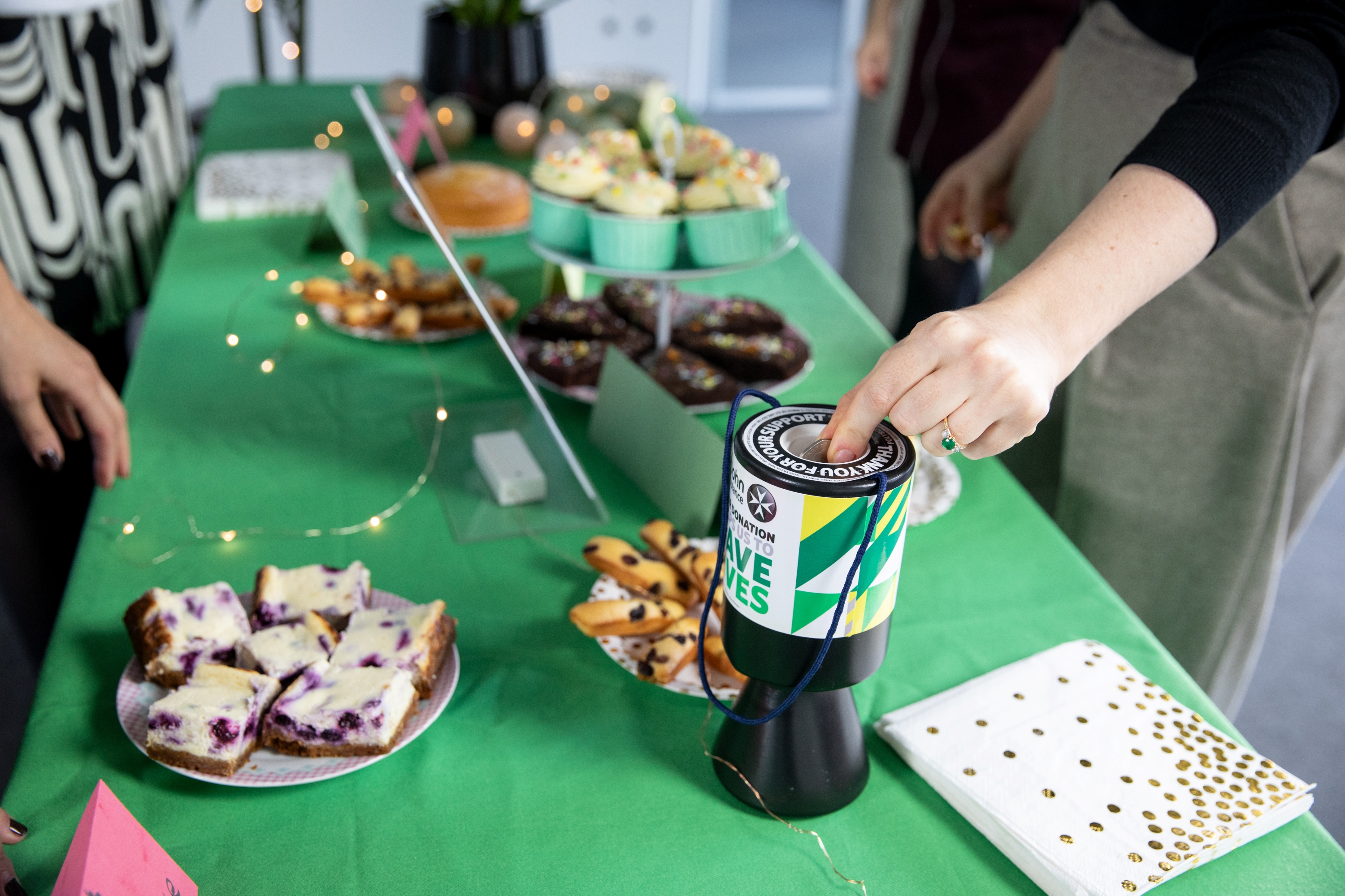 A photograph of a bake sale. A variety of cakes and baked goods sit on top of a table covered by a St John tablecloth. In the foreground is a St John Ambulance donation box, with someone dropping money into it.