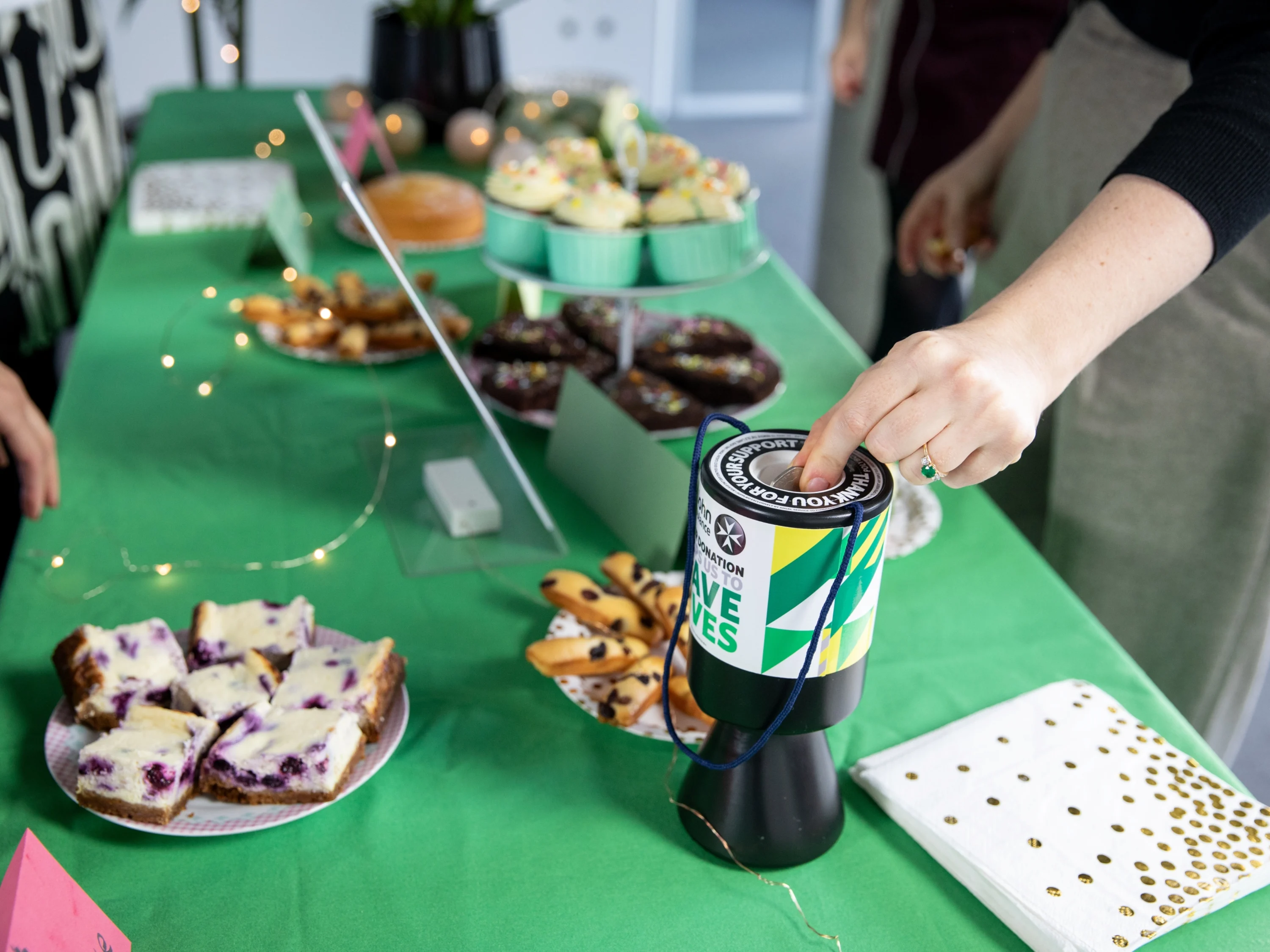 A photograph of a bake sale. A variety of cakes and baked goods sit on top of a table covered by a St John tablecloth. In the foreground is a St John Ambulance donation box, with someone dropping money into it.