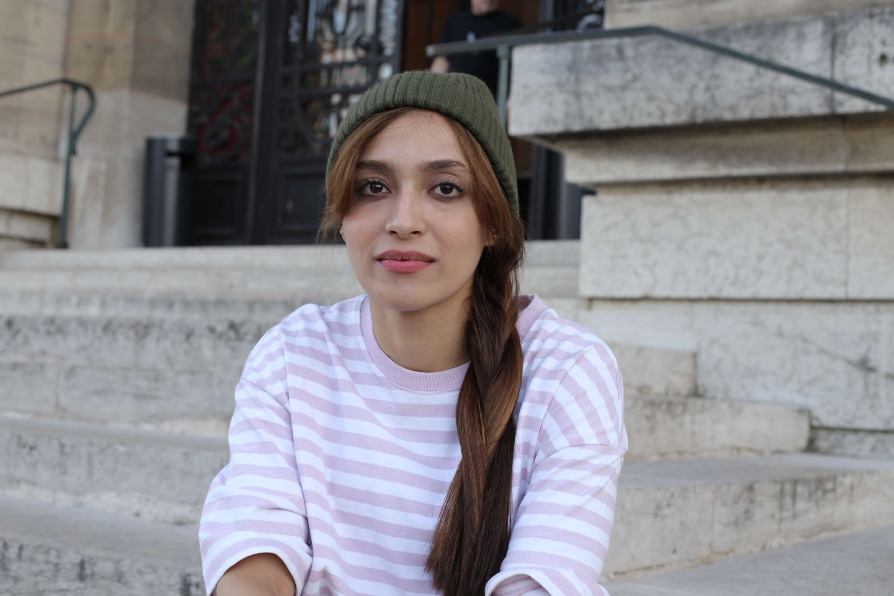 A photograph of Kate* sitting down and staring at the camera. She is wearing a pink, stripey top and a green beanie.