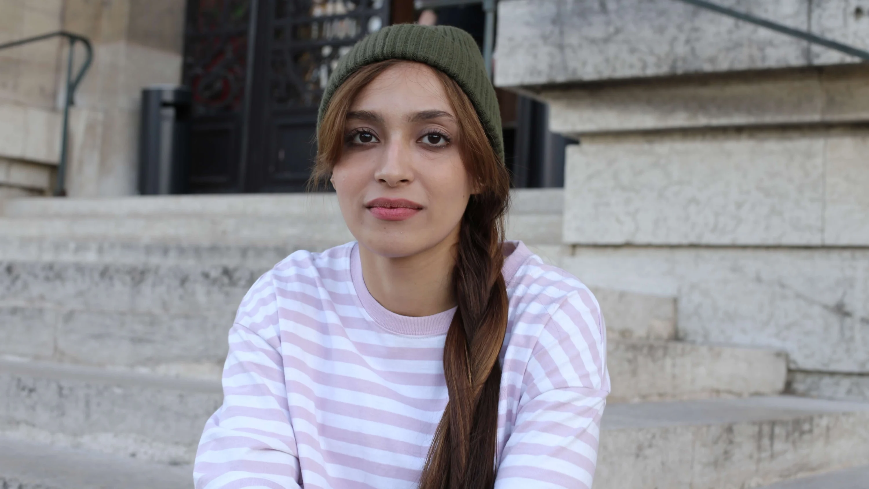 A photograph of Kate* sitting down and staring at the camera. She is wearing a pink, stripey top and a green beanie.