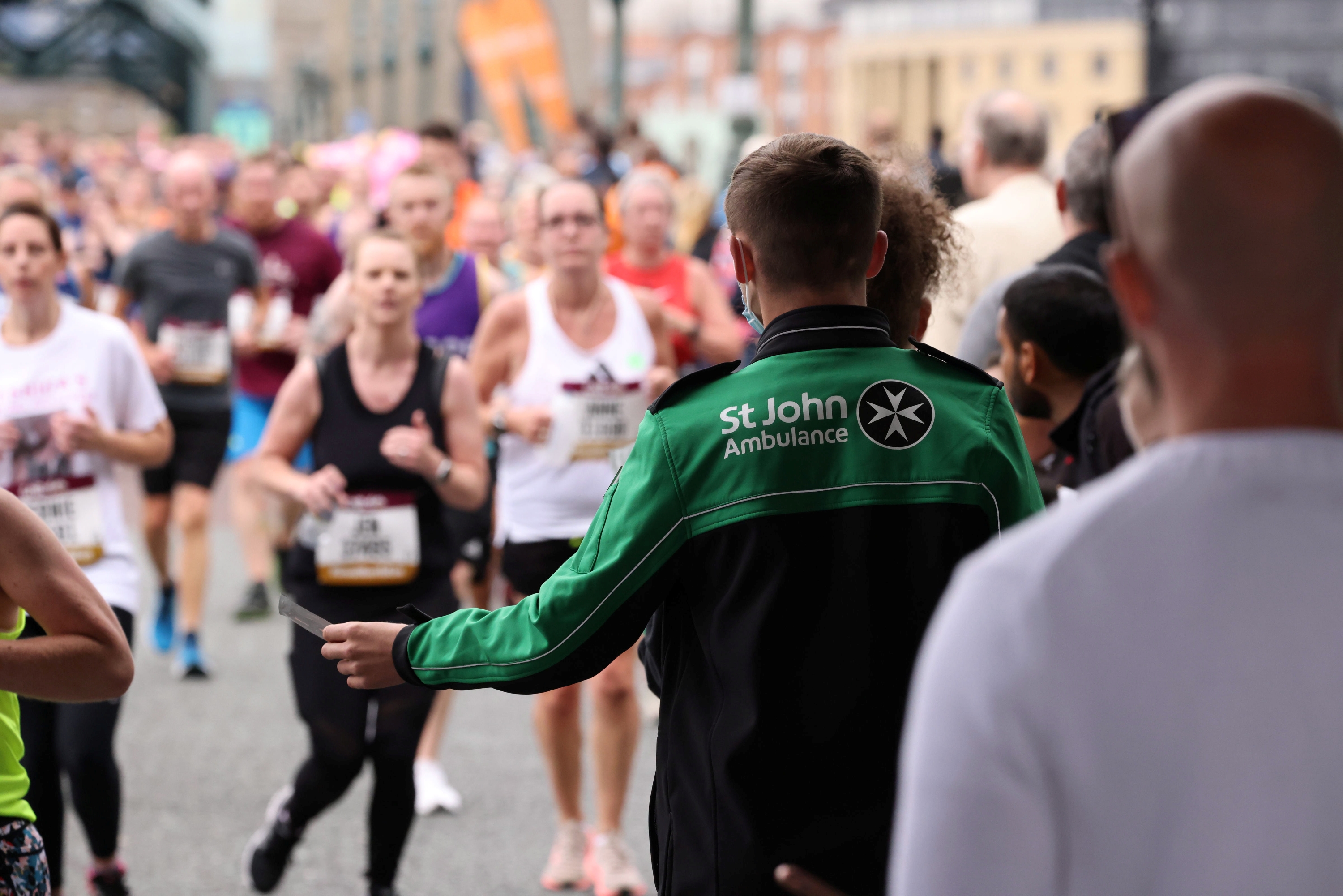 The back of a St John Ambulance volunteer at a race who is holding out a small piece of paper. There is a large group of runners running towards the volunteer and other people. 