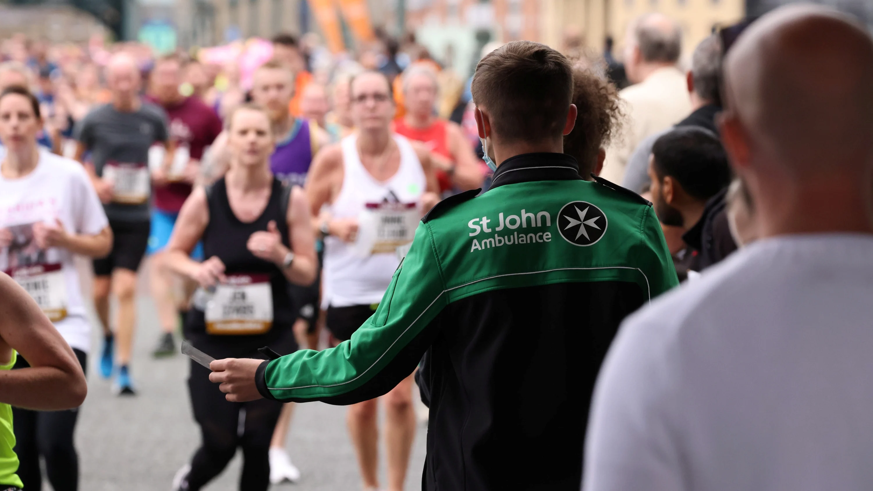 The back of a St John Ambulance volunteer at a race who is holding out a small piece of paper. There is a large group of runners running towards the volunteer and other people.