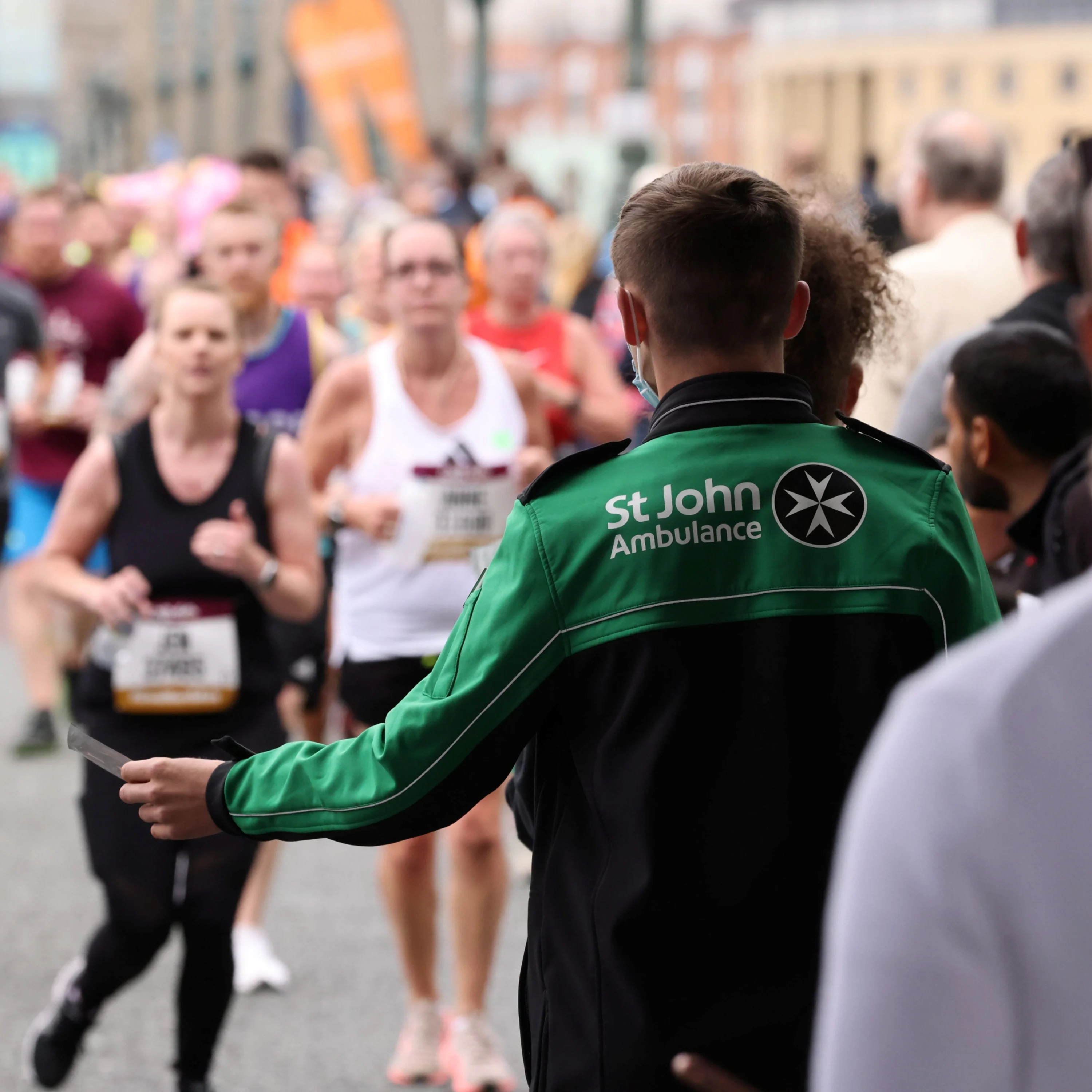 The back of a St John Ambulance volunteer at a race who is holding out a small piece of paper. There is a large group of runners running towards the volunteer and other people.
