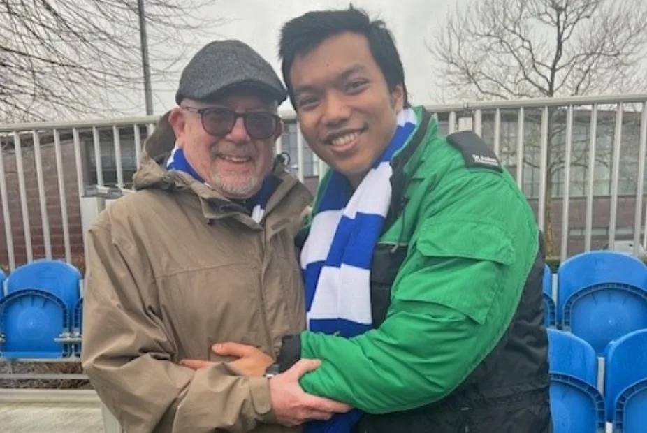 Paul Archer and Prince Tandukar by the seats at Chelsea Football Club, both smiling and standing with their arms around each other 