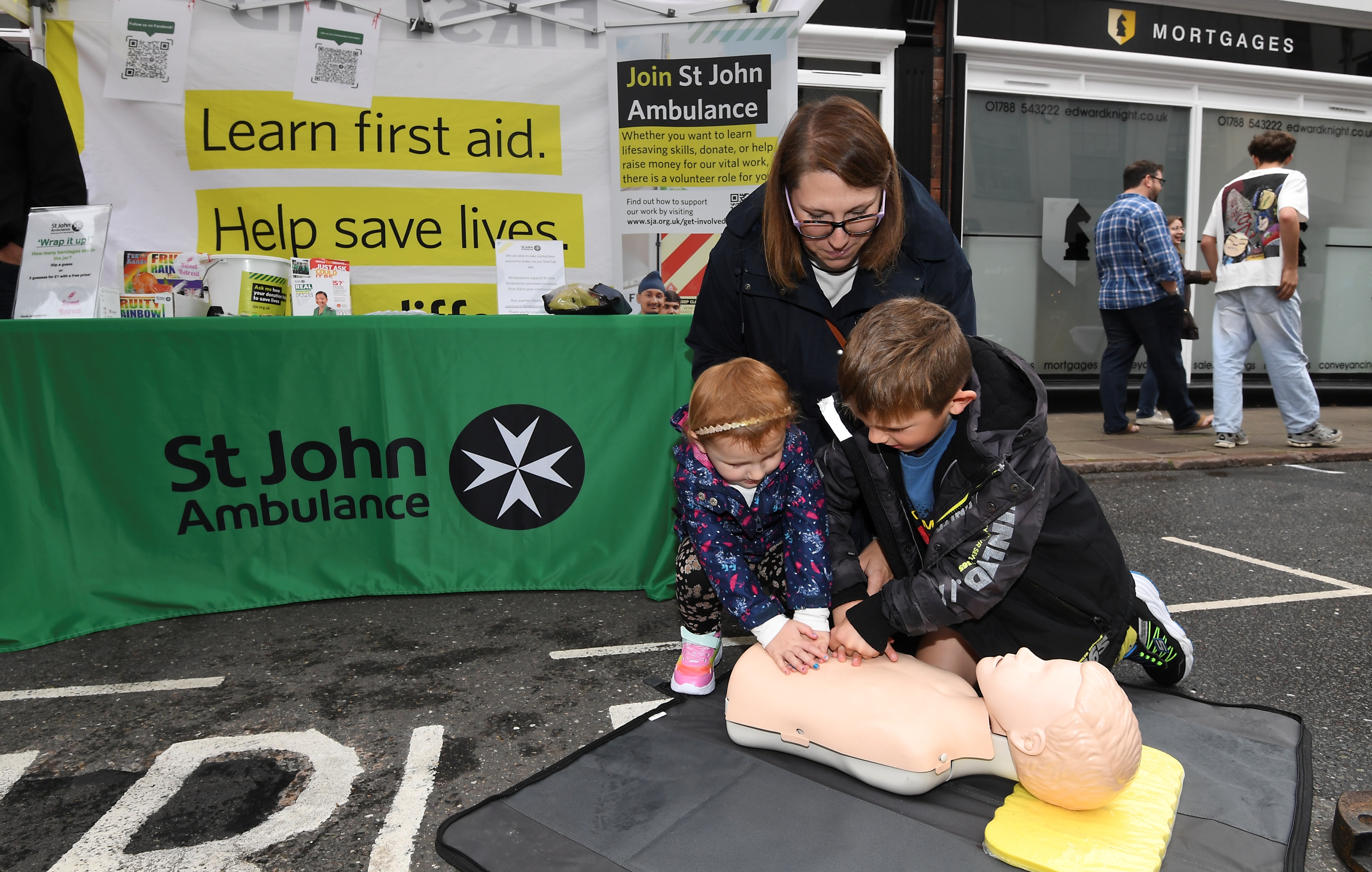Photograph of a St John community demonstration. Two small children, a boy and a girl, perform chest compressions on a Manikin as their mother watches.