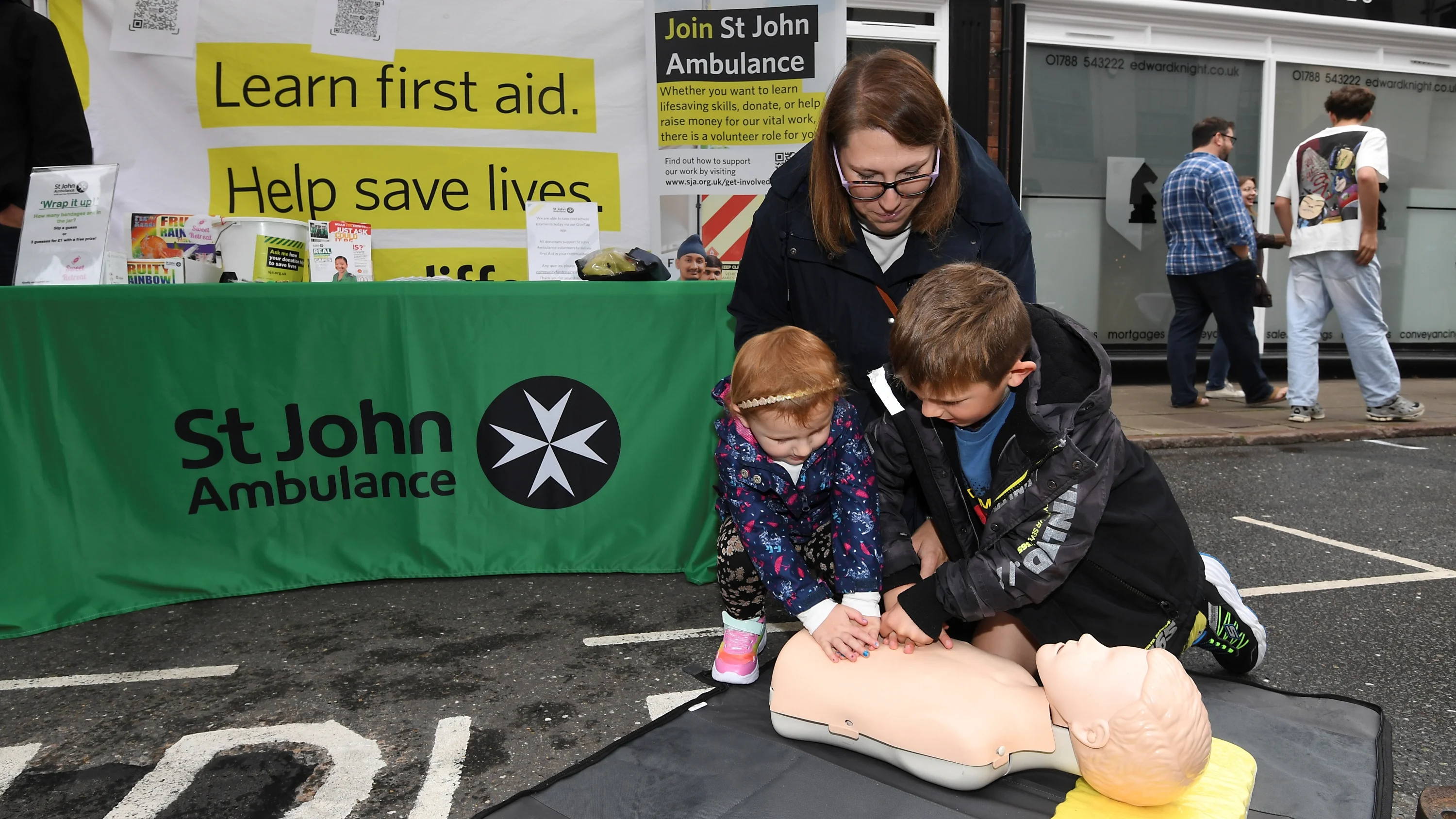 Photograph of a St John community demonstration. Two small children, a boy and a girl, perform chest compressions on a Manikin as their mother watches.