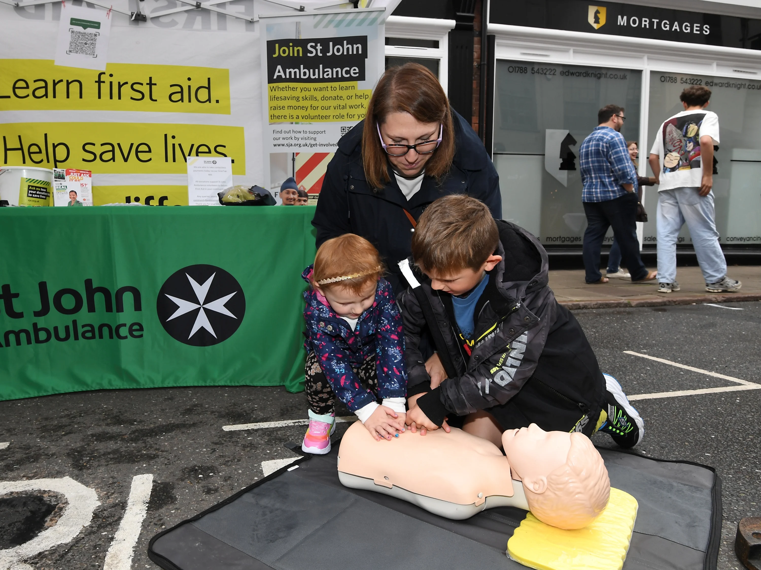 Photograph of a St John community demonstration. Two small children, a boy and a girl, perform chest compressions on a Manikin as their mother watches.