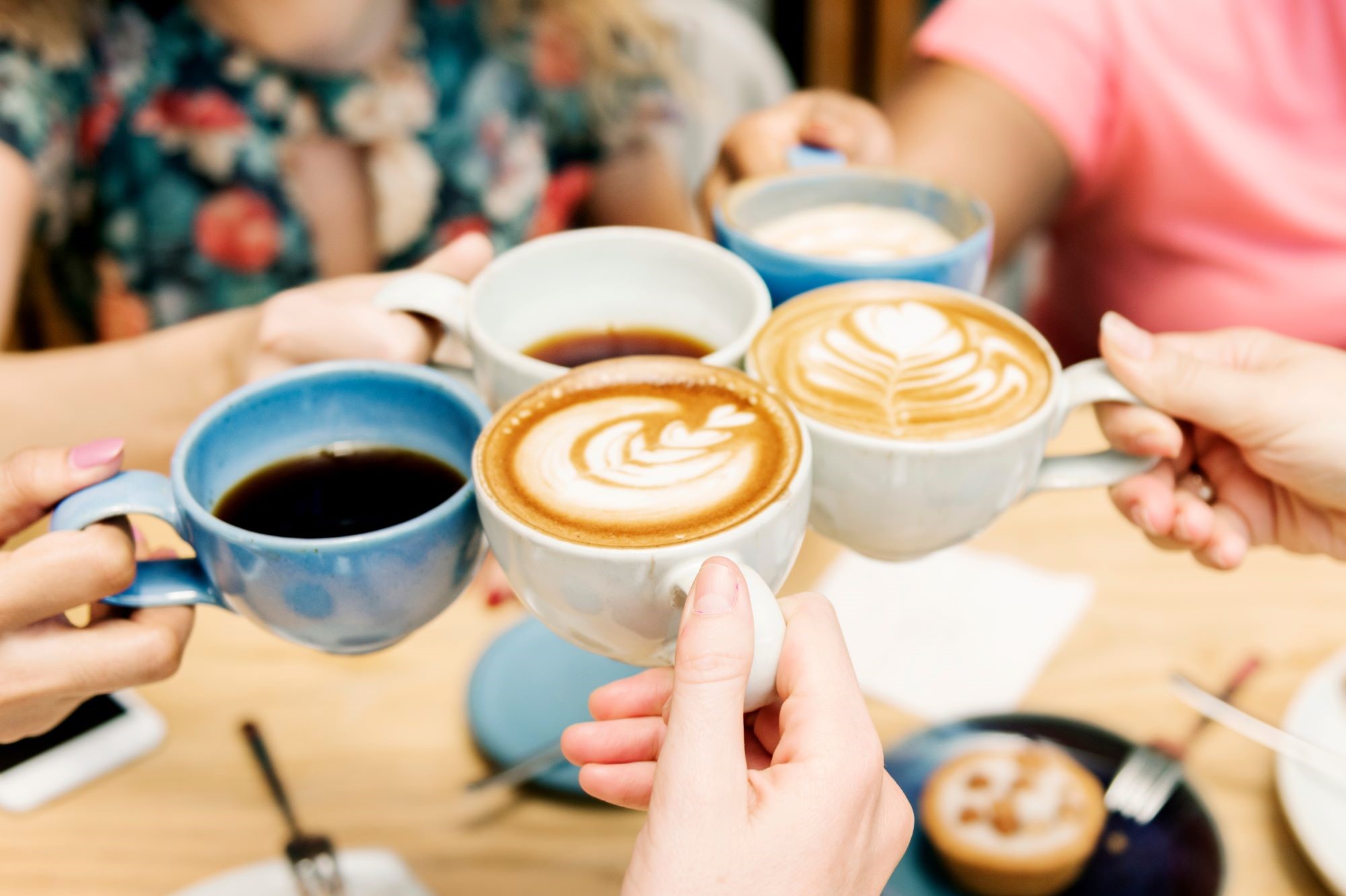 Five people holding coffee and tea cups doing a 'cheers' above a table covered in plates, cutlery, and cakes. 