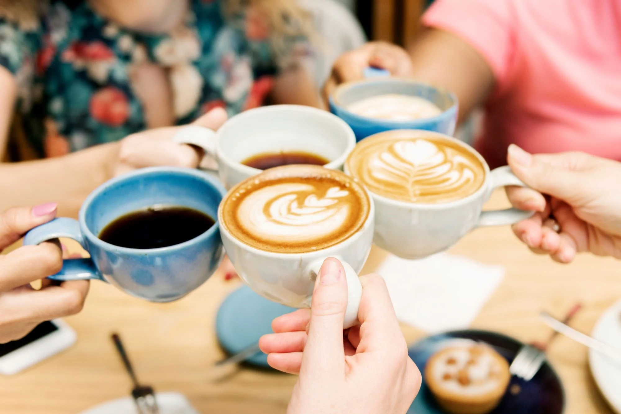 Five people holding coffee and tea cups doing a 'cheers' above a table covered in plates, cutlery, and cakes.