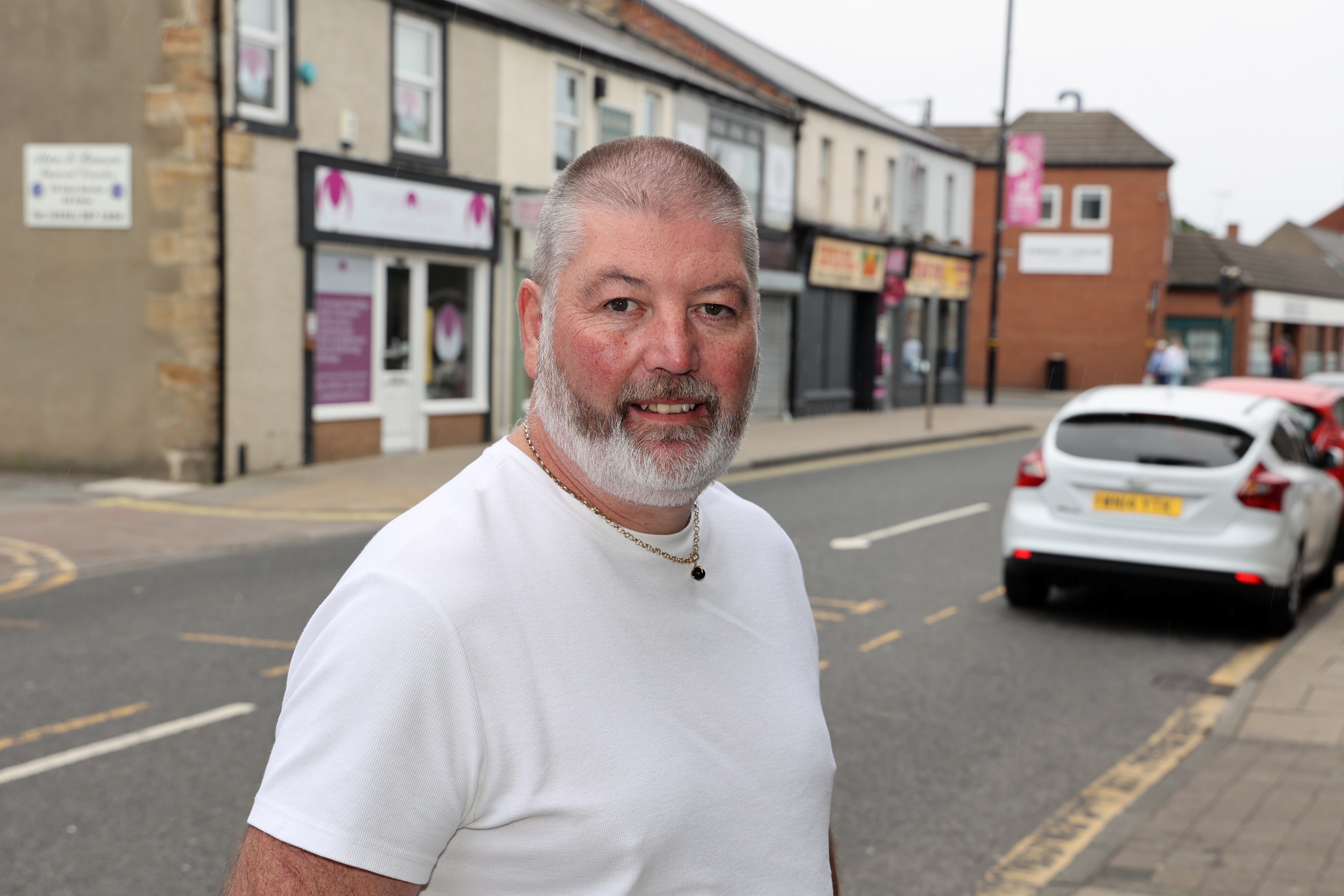 A photograph of Peter Batey in a white t-shirt, standing in the street and smiling at the camera.