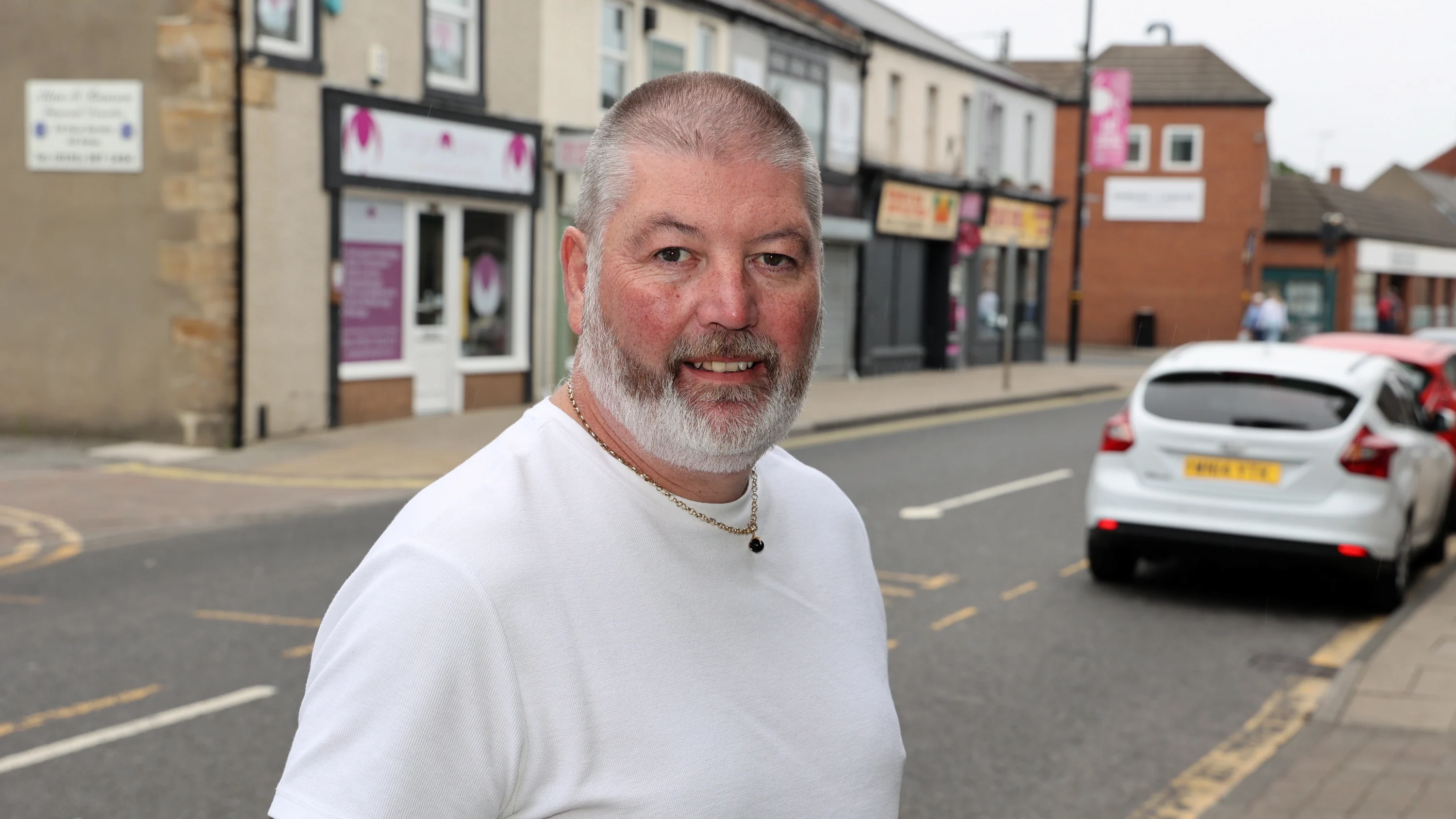 A photograph of Peter Batey in a white t-shirt, standing in the street and smiling at the camera.