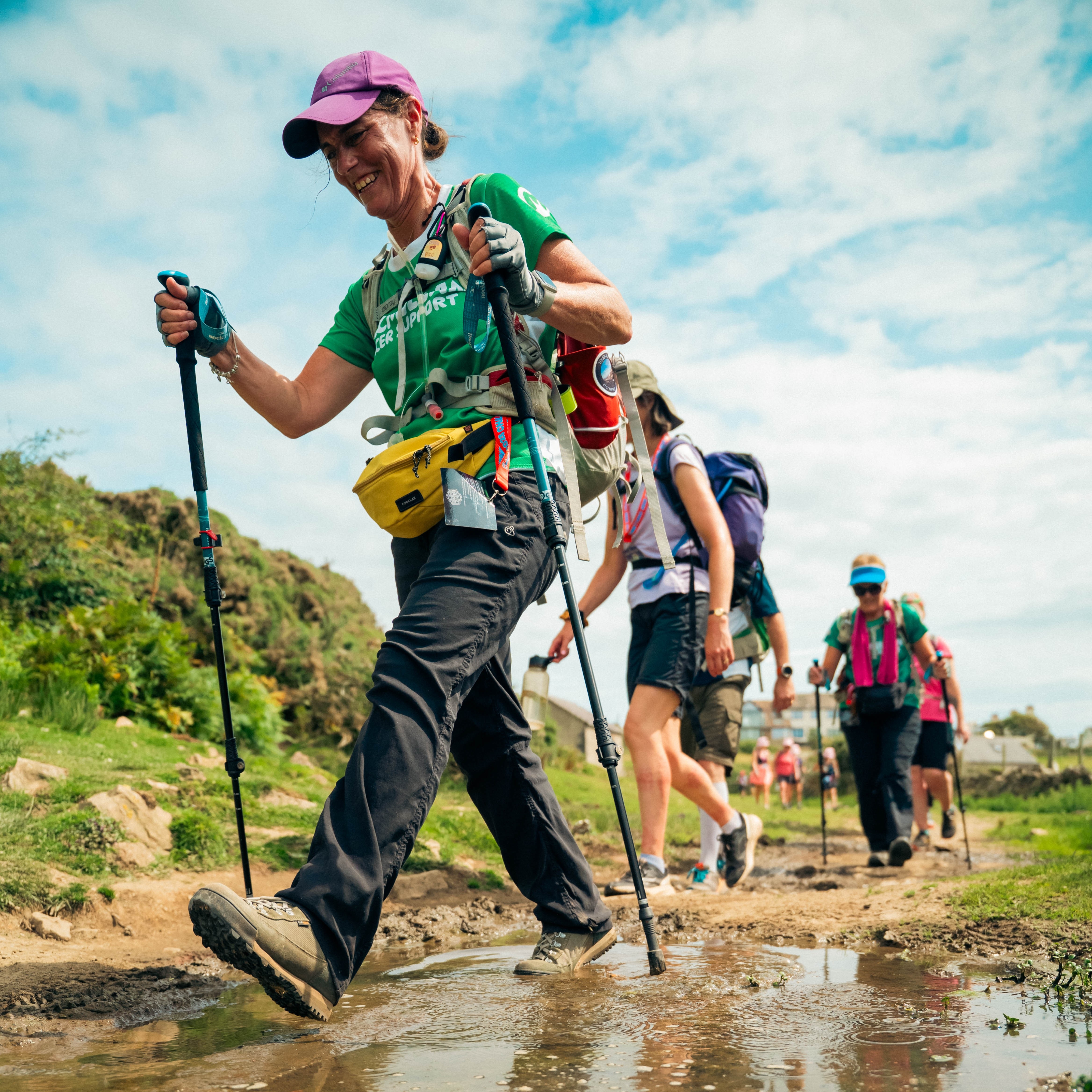 A group of people in hiking gear and using walking sticks walking along a very muddy and wet footpath.