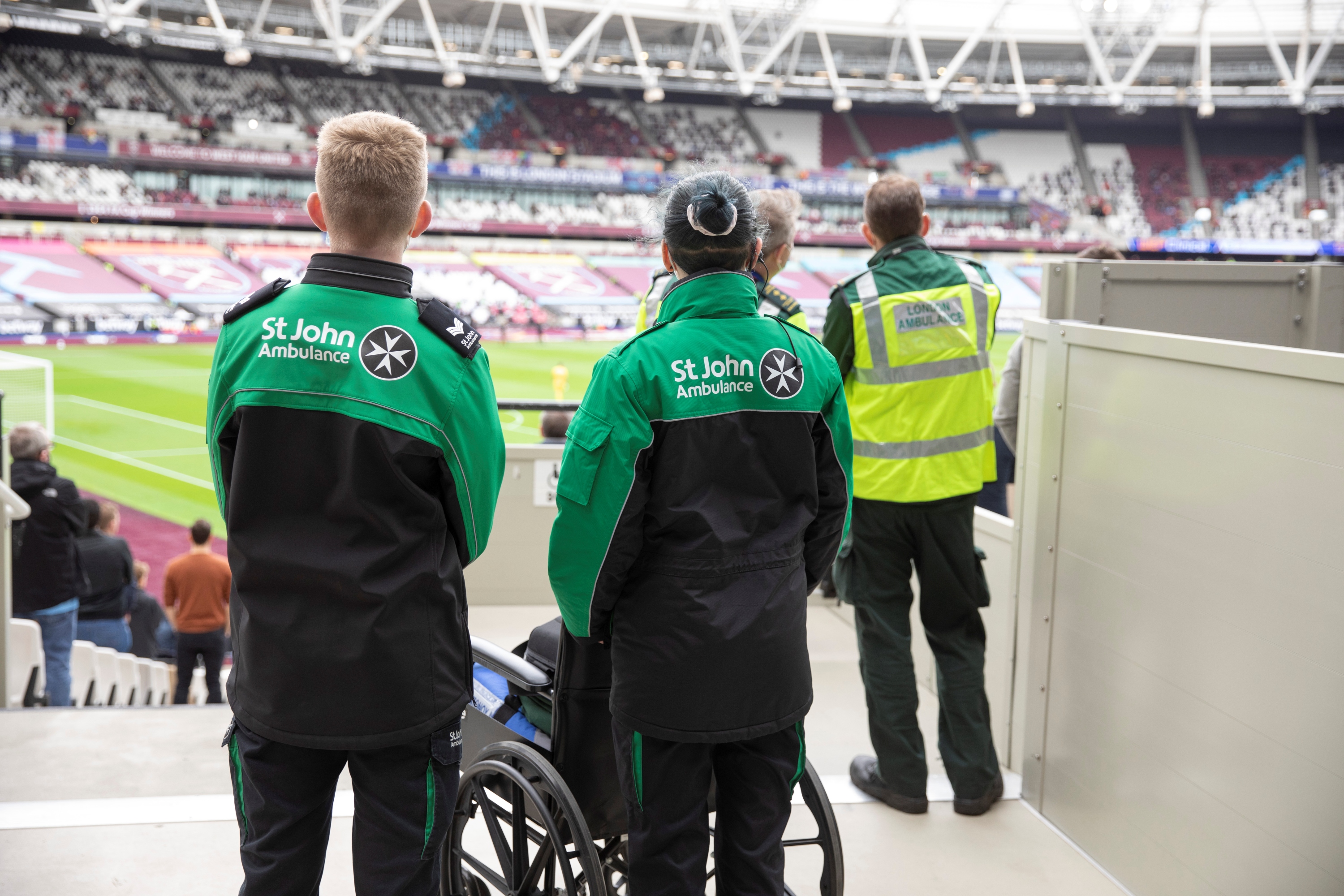 The backs of three St John Ambulance volunteers stood at the side of a large sports stadium looking out to the pitch. In front of one of them is a wheelchair. 