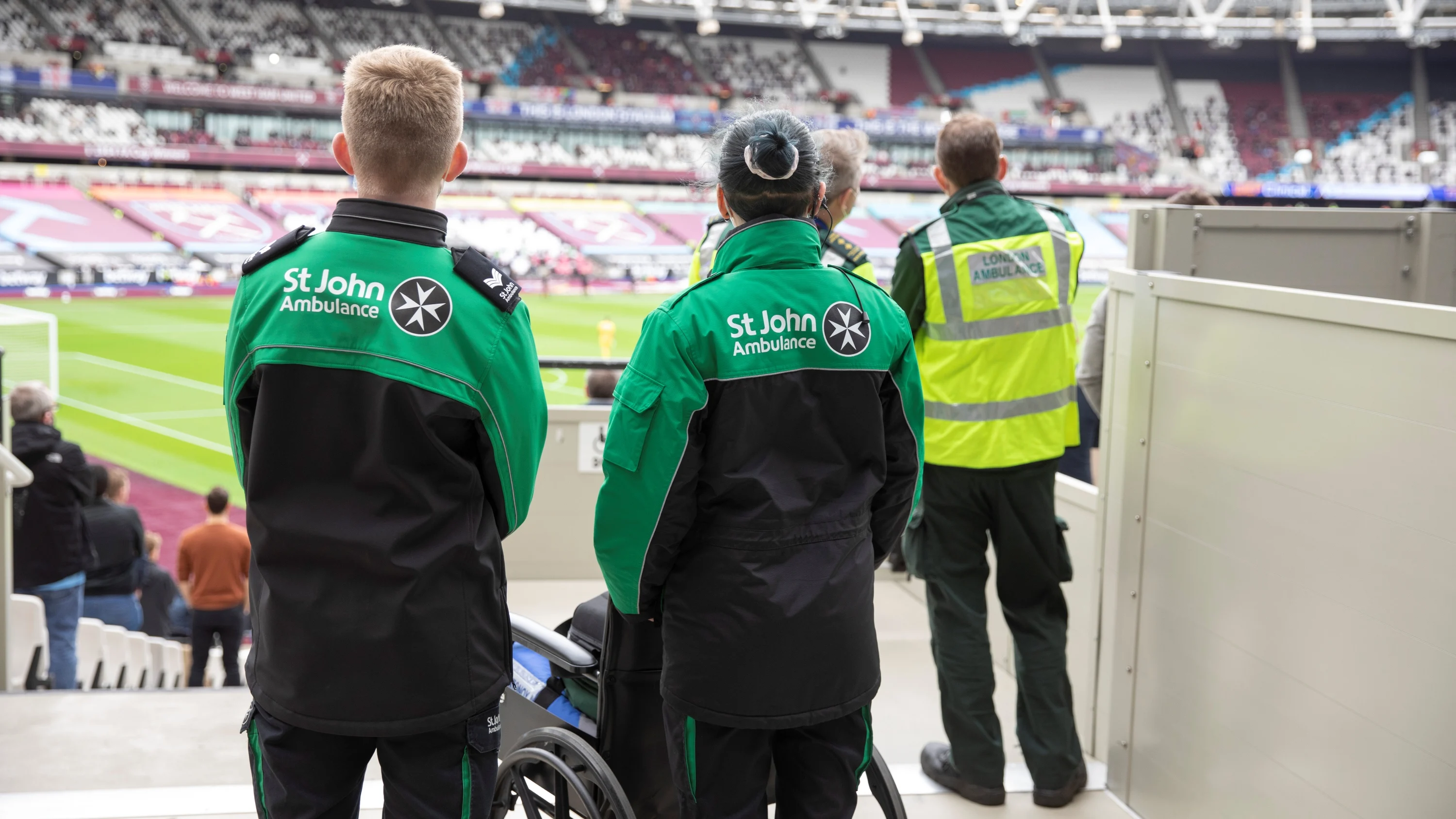 The backs of three St John Ambulance volunteers stood at the side of a large sports stadium looking out to the pitch. In front of one of them is a wheelchair.