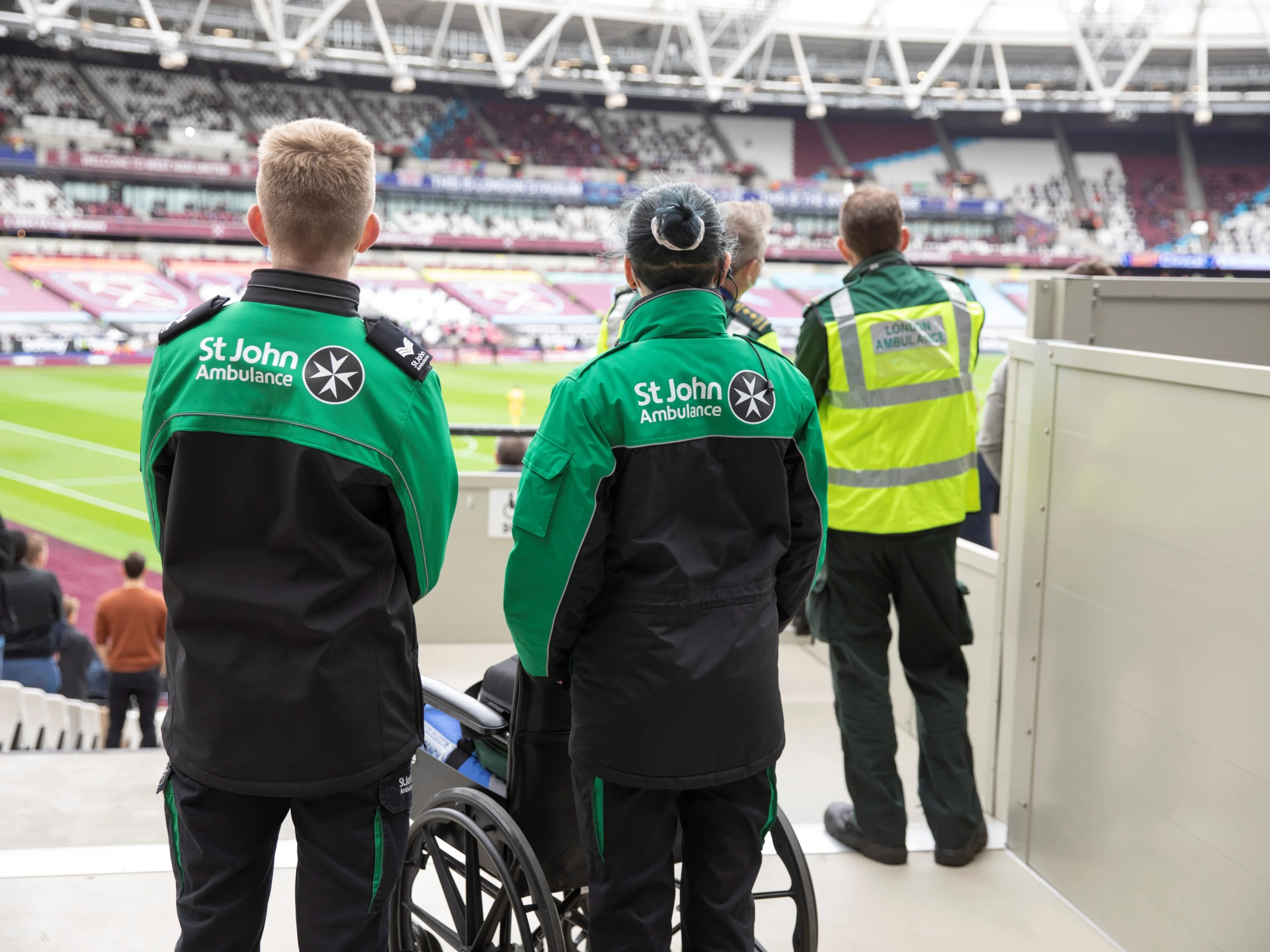 The backs of three St John Ambulance volunteers stood at the side of a large sports stadium looking out to the pitch. In front of one of them is a wheelchair.