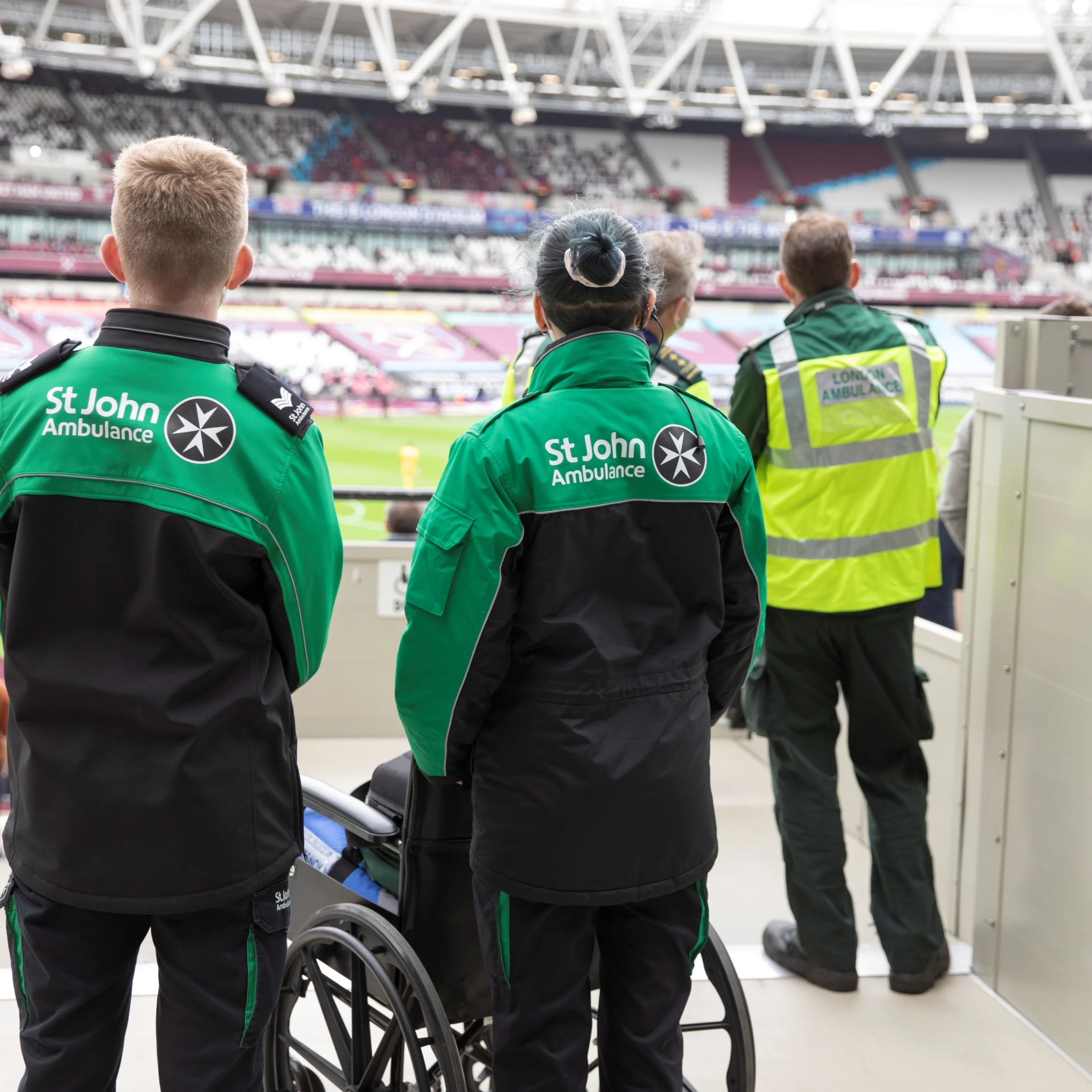 The backs of three St John Ambulance volunteers stood at the side of a large sports stadium looking out to the pitch. In front of one of them is a wheelchair.