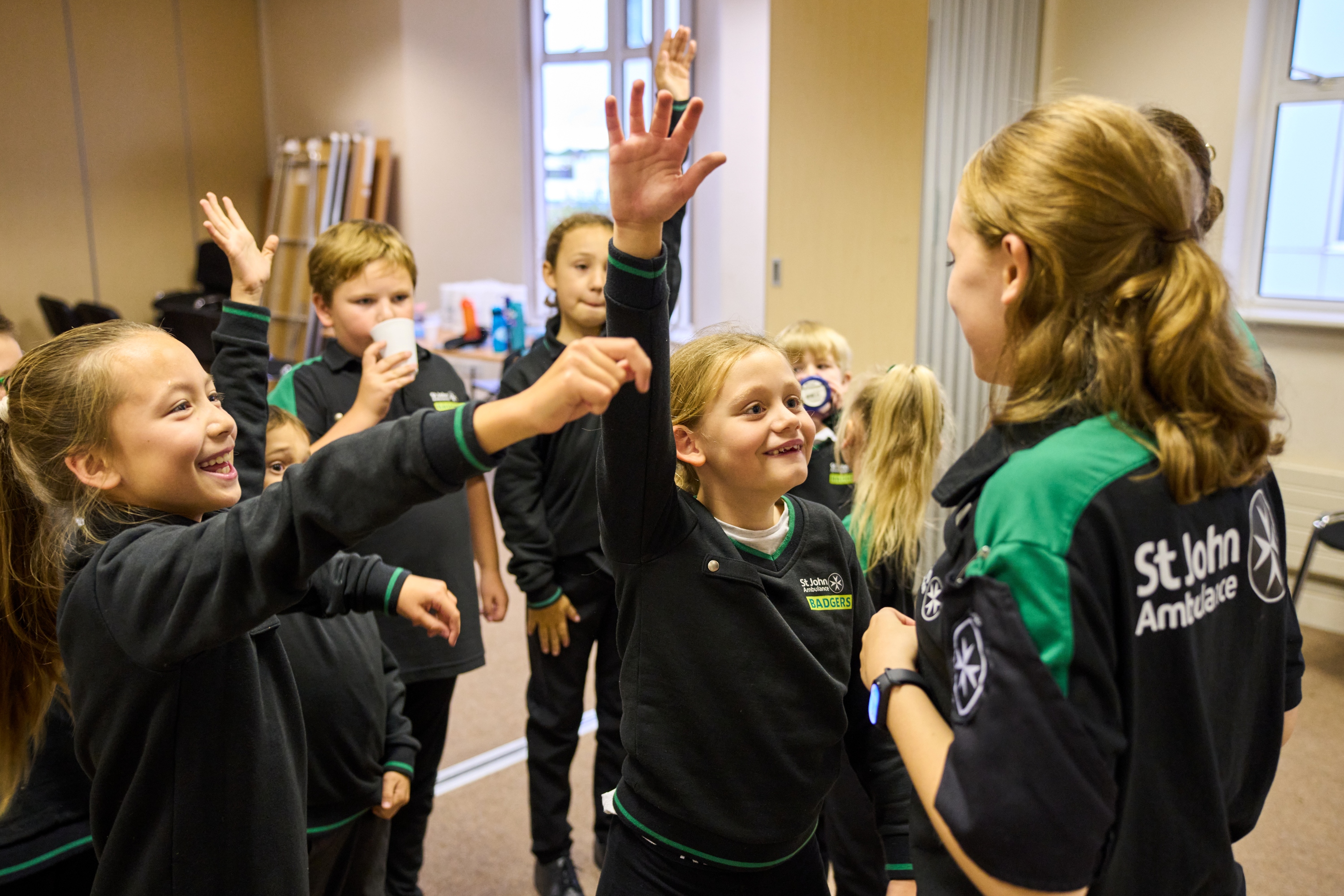 A group of Badgers inside standing in front of a Youth Leader smiling. Some of the Badgers have their hands up.