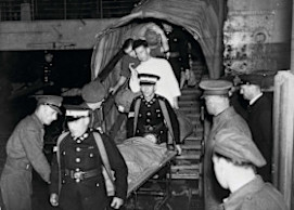 A black and white photograph of St John volunteers carrying a patient on a stretcher. They are surrounded by men in uniform.