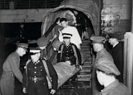 A black and white photograph of St John volunteers carrying a patient on a stretcher. They are surrounded by men in uniform.