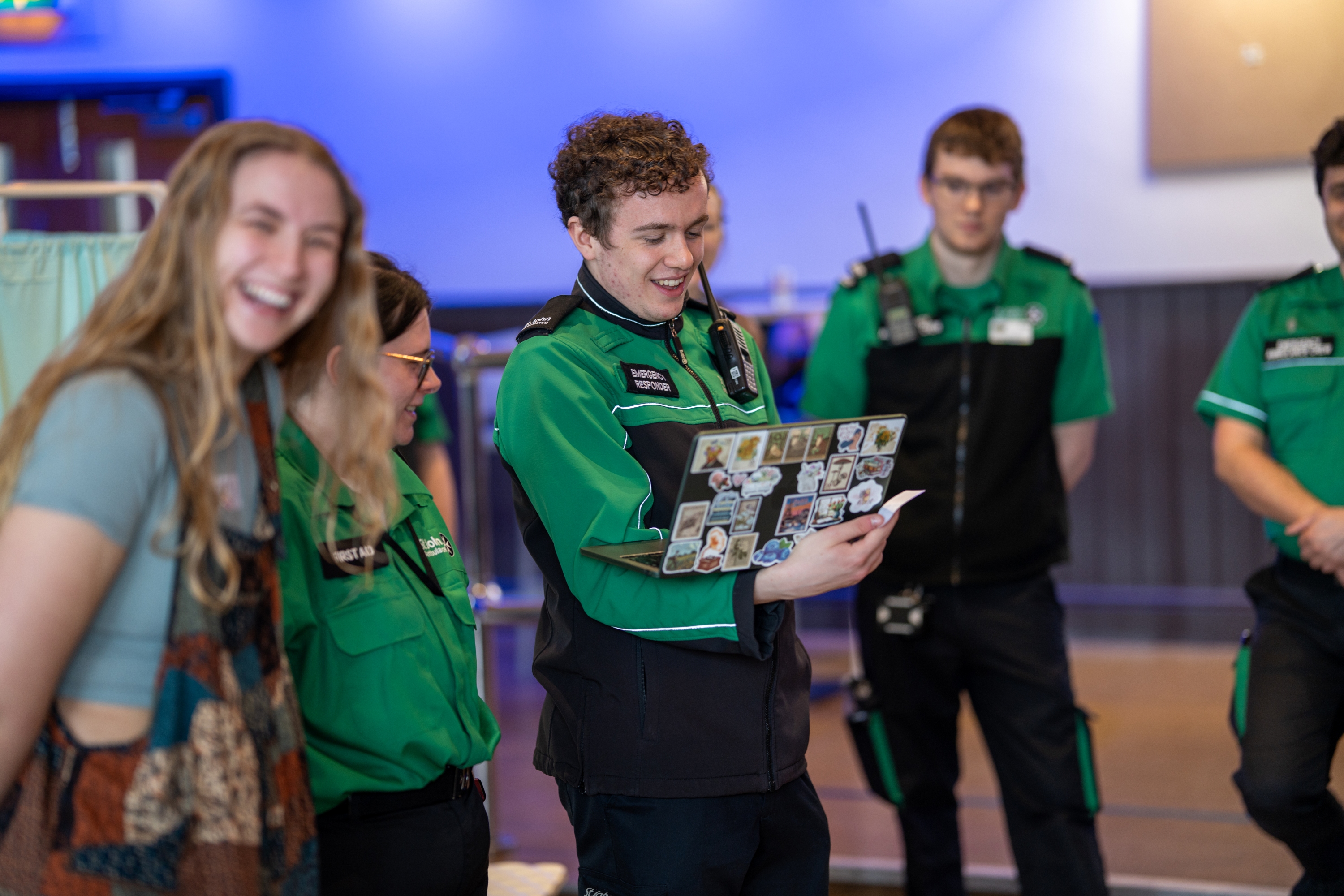 Five students standing in a group in a hall. Four are wearing St John Ambulance volunteer uniforms. One of them is holding a laptop. 