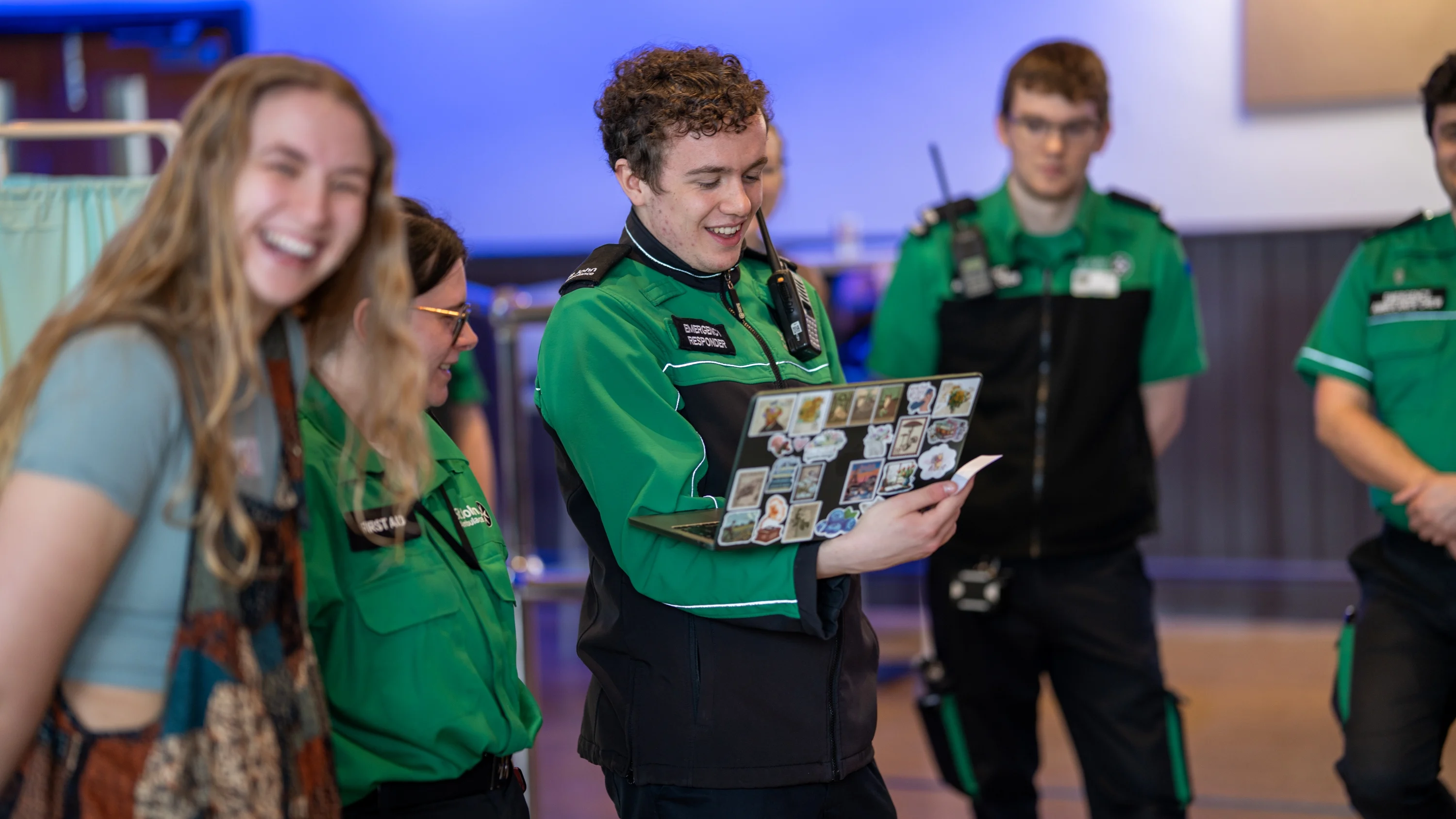 Five students standing in a group in a hall. Four are wearing St John Ambulance volunteer uniforms. One of them is holding a laptop.