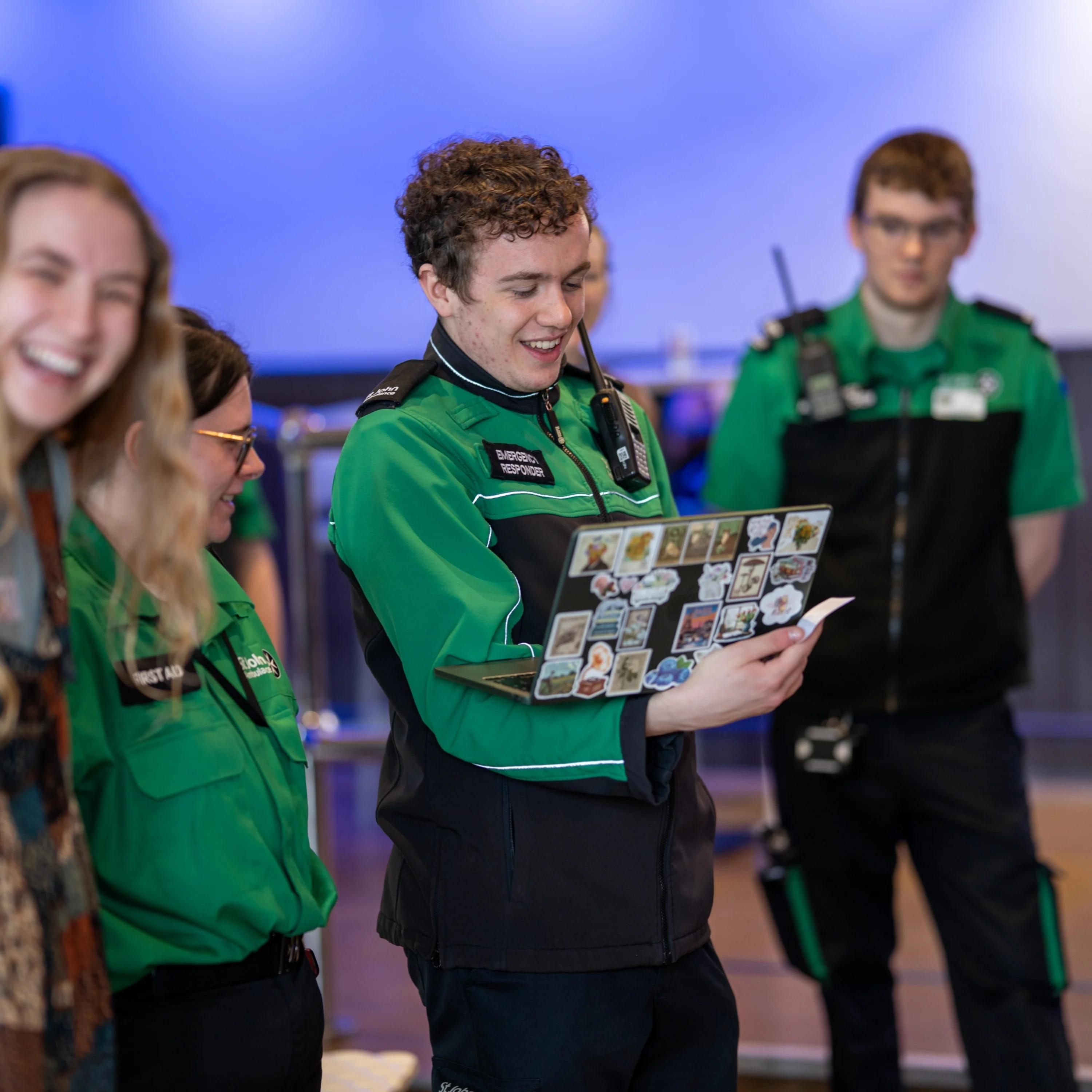 Five students standing in a group in a hall. Four are wearing St John Ambulance volunteer uniforms. One of them is holding a laptop.