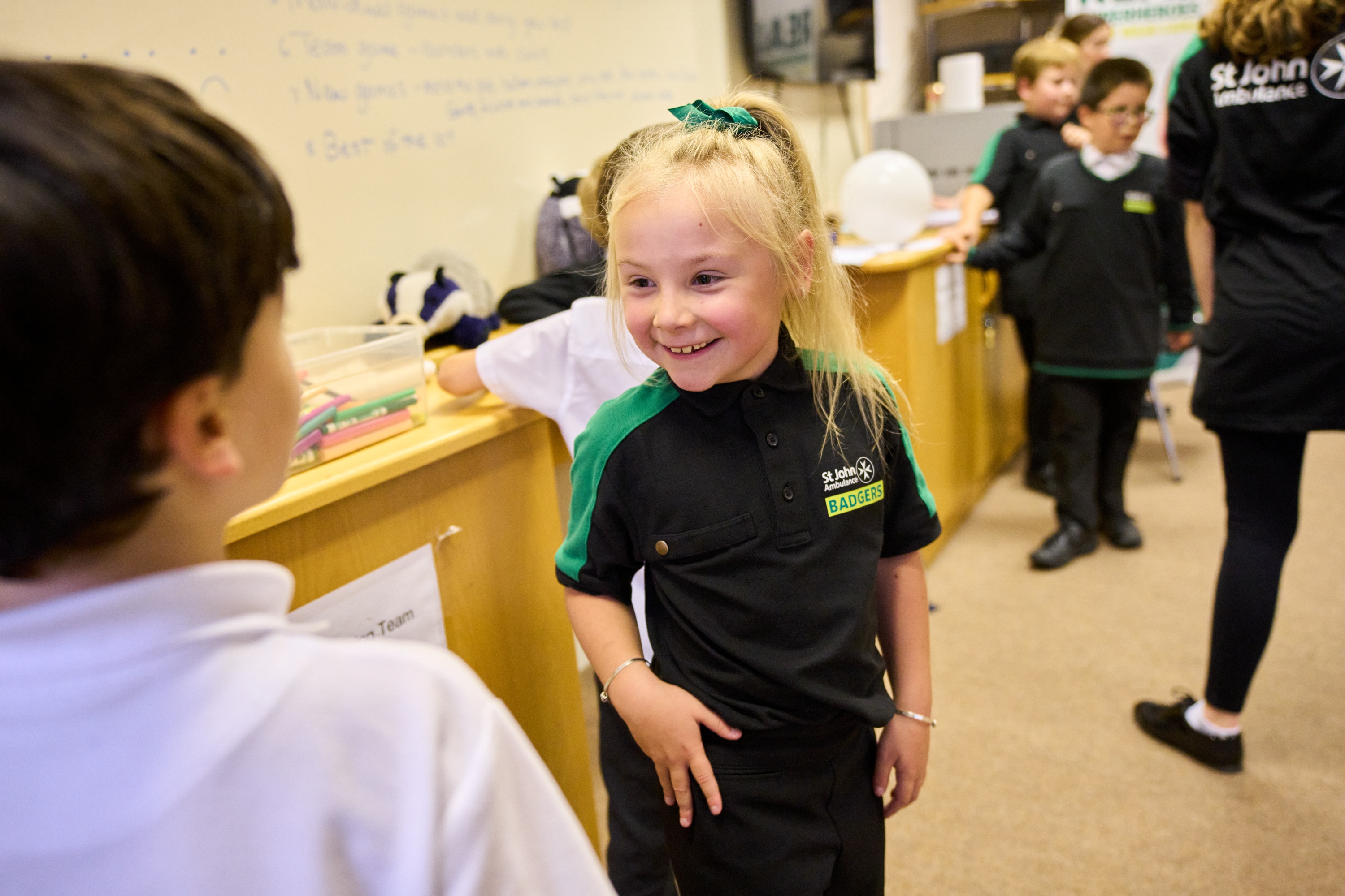 A Badger standing and smiling in front of another Badger with others standing around in the background. Behind them, there is a whiteboard on the wall with writing all over it and boxes of pencils and a toy badger. 