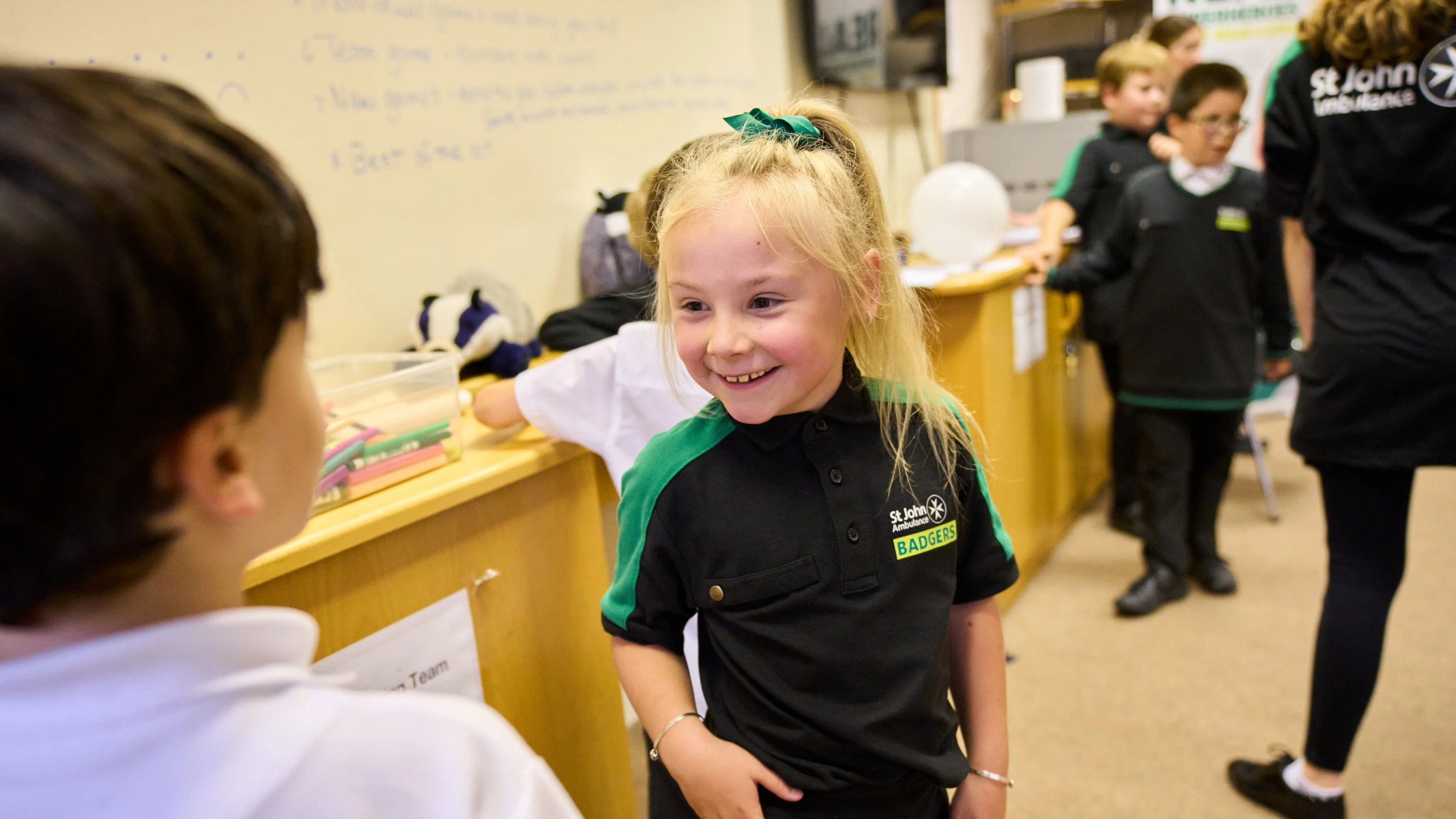 A Badger standing and smiling in front of another Badger with others standing around in the background. Behind them, there is a whiteboard on the wall with writing all over it and boxes of pencils and a toy badger.