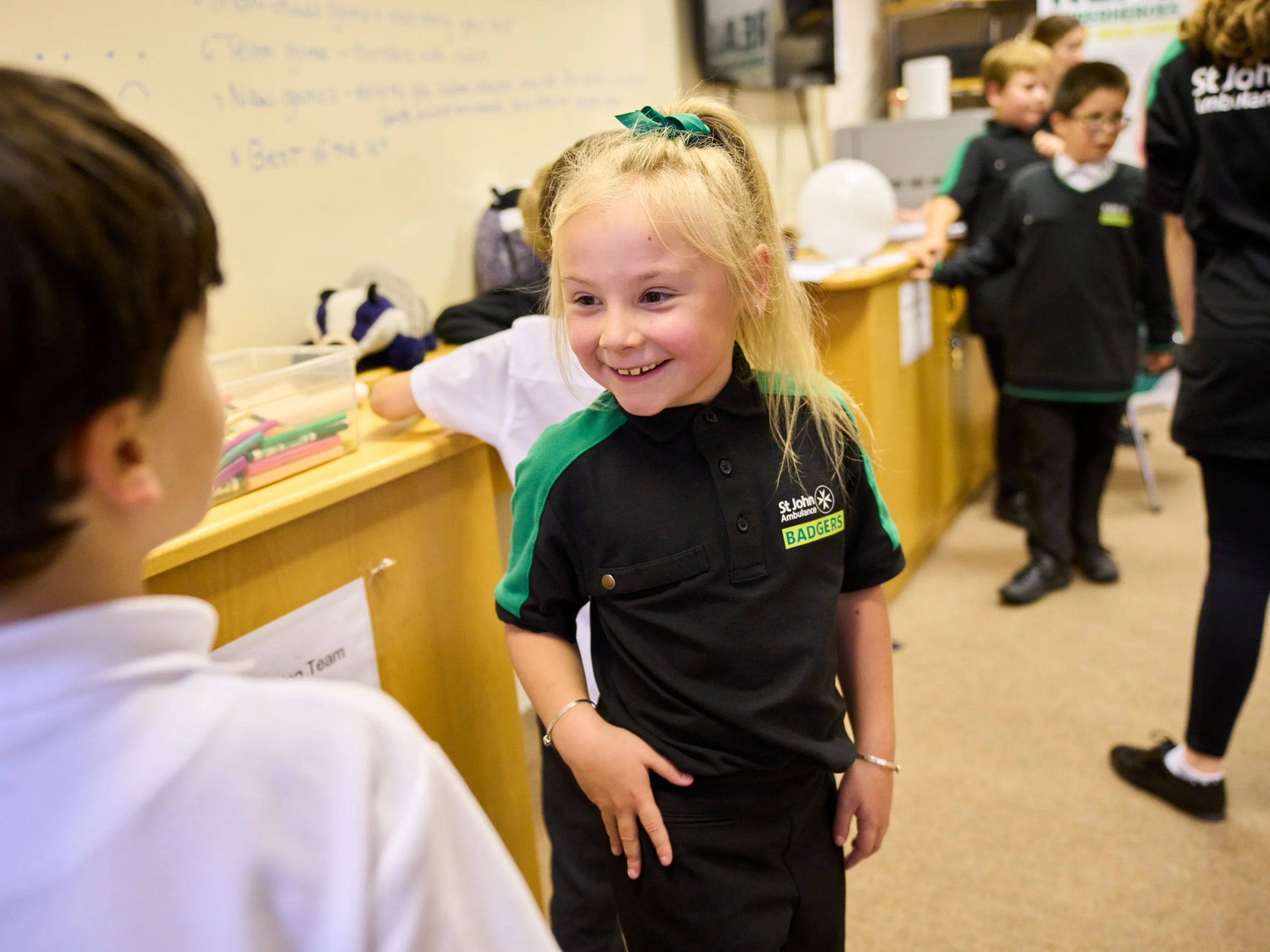 A Badger standing and smiling in front of another Badger with others standing around in the background. Behind them, there is a whiteboard on the wall with writing all over it and boxes of pencils and a toy badger.