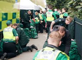 A photograph of SJA volunteers working with NHS paramedics. Ambulances and medical tents can be seen in the background.