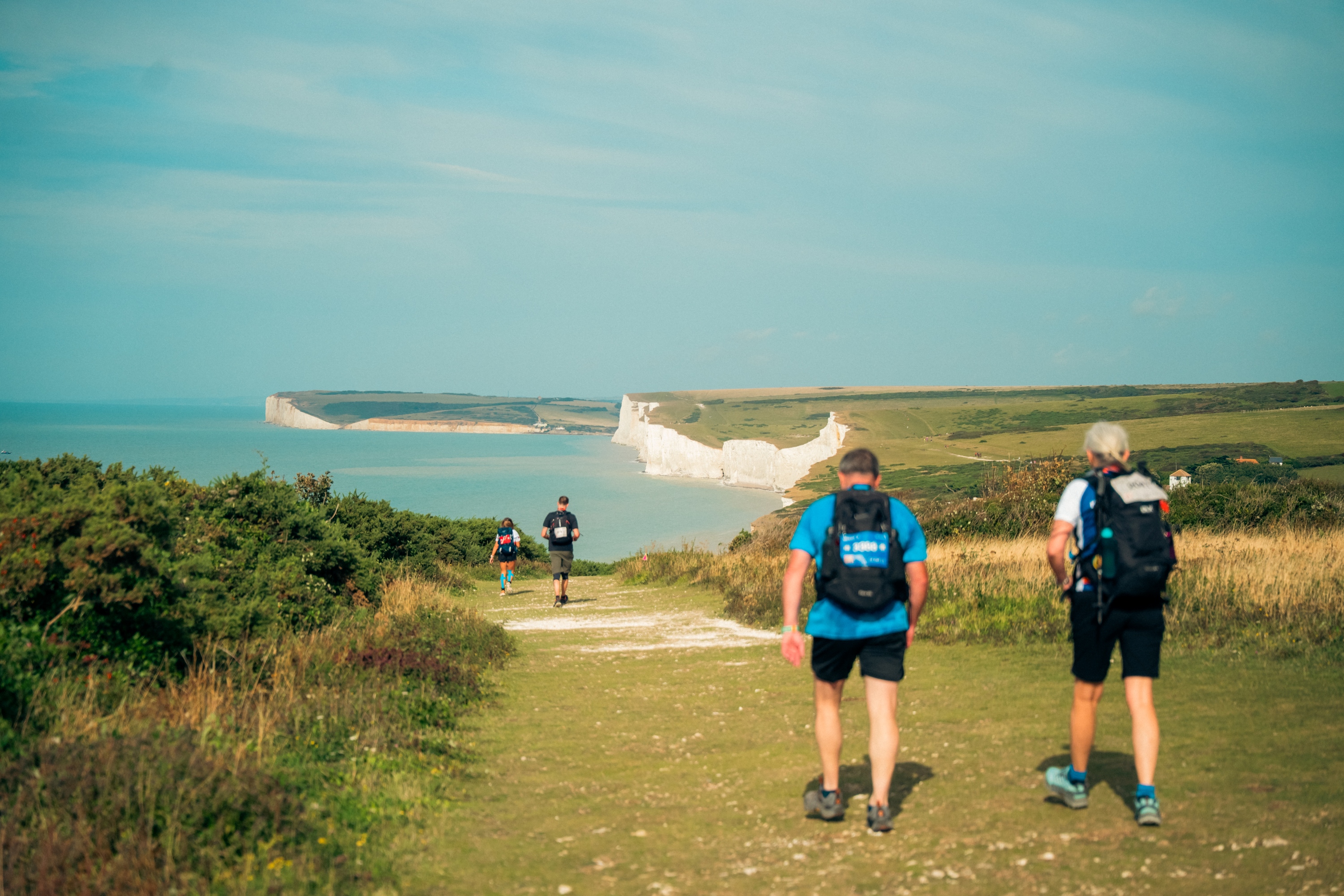 Four people walking along the South Downs coastal path. In the distance are white cliffs and the sea.