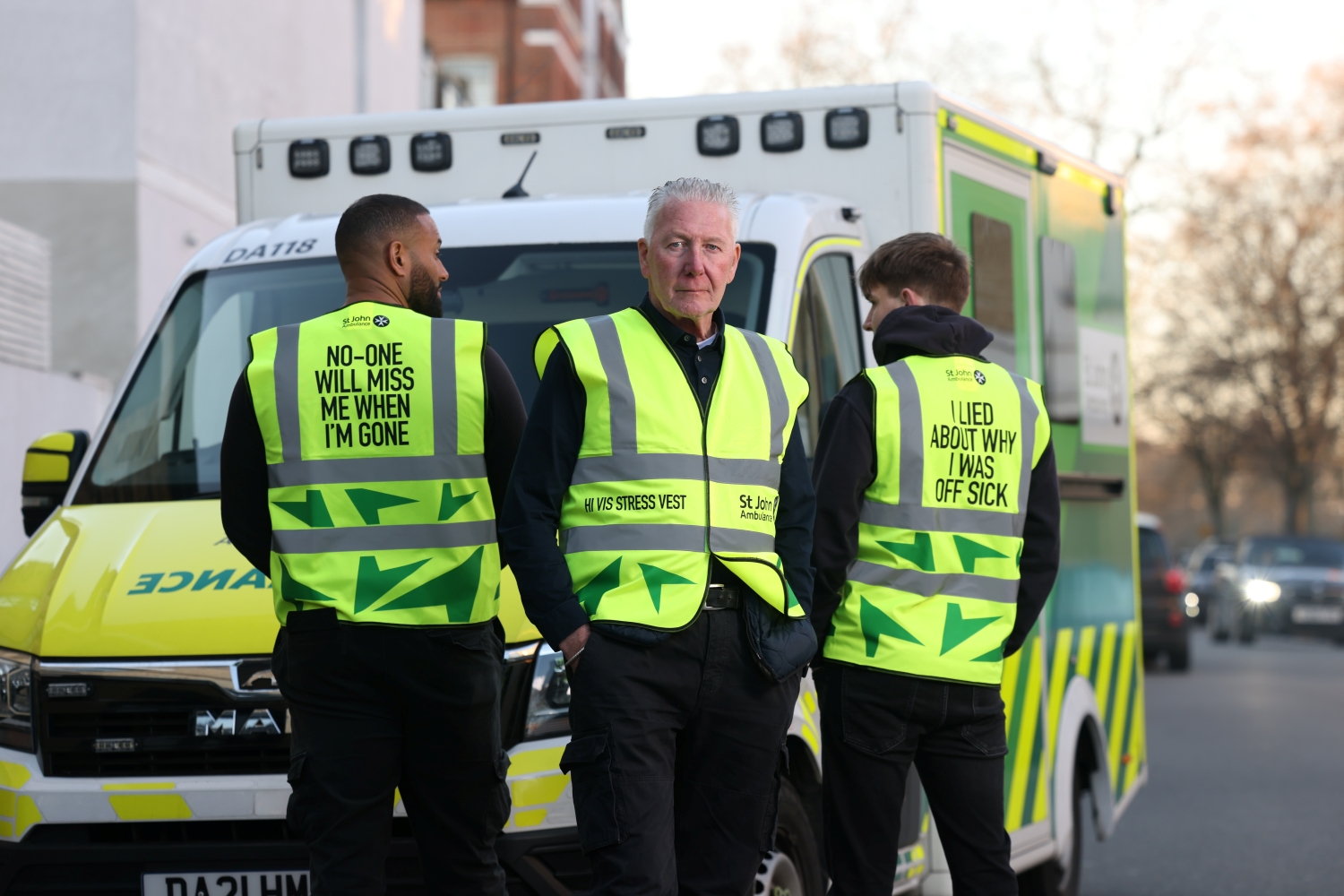 Three people wearing the Hi Vis Stress Vest and stood in front of an ambulance. On the back of one vest it says 'No-one will miss me when I'm gone' and another vest says 'I lied about why I was off sick'.