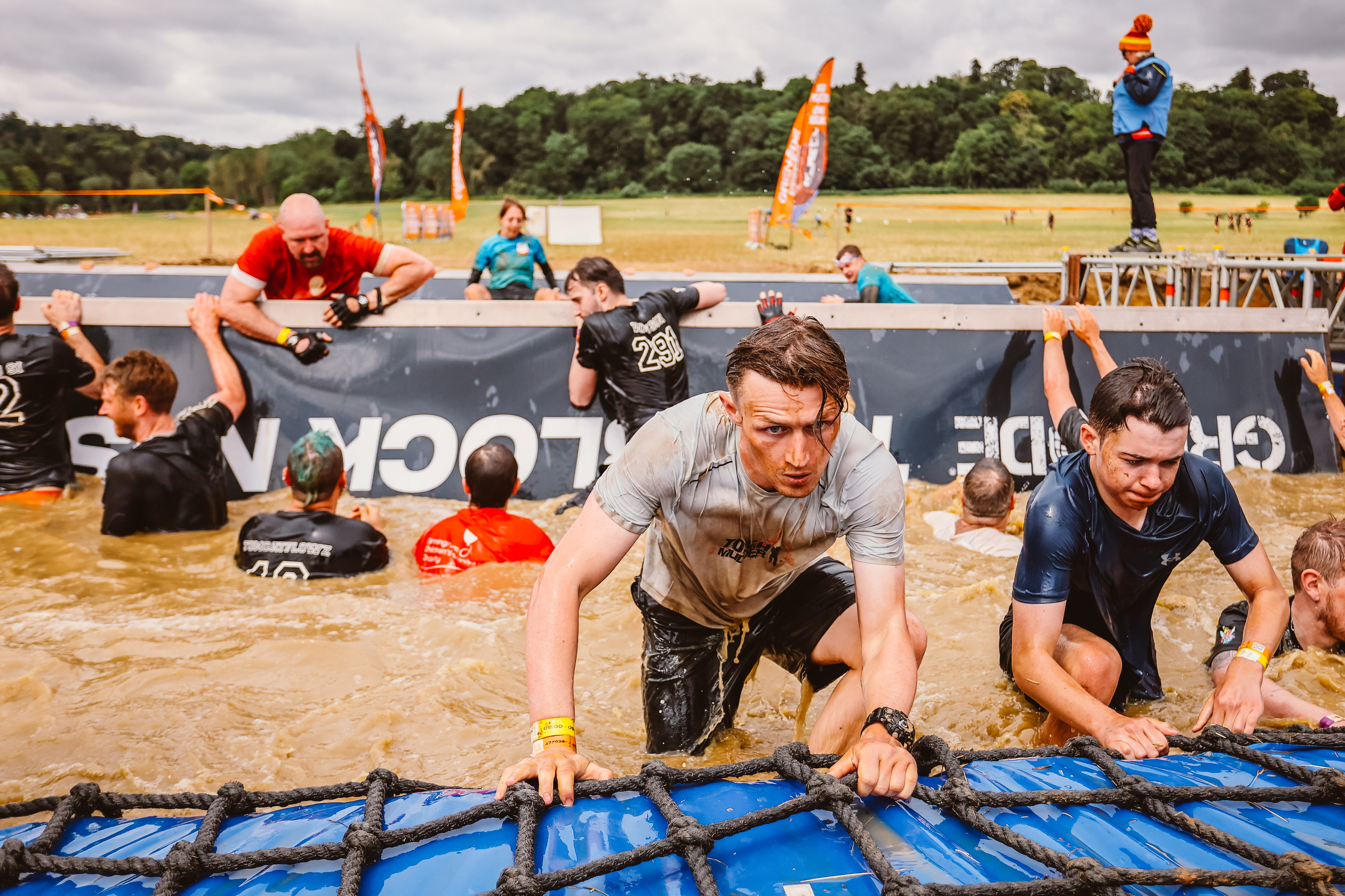 A Tough Mudder event outside in a field. A group of people are in a large pool of muddy water, some are climbing out of the sides holding onto ropes. 