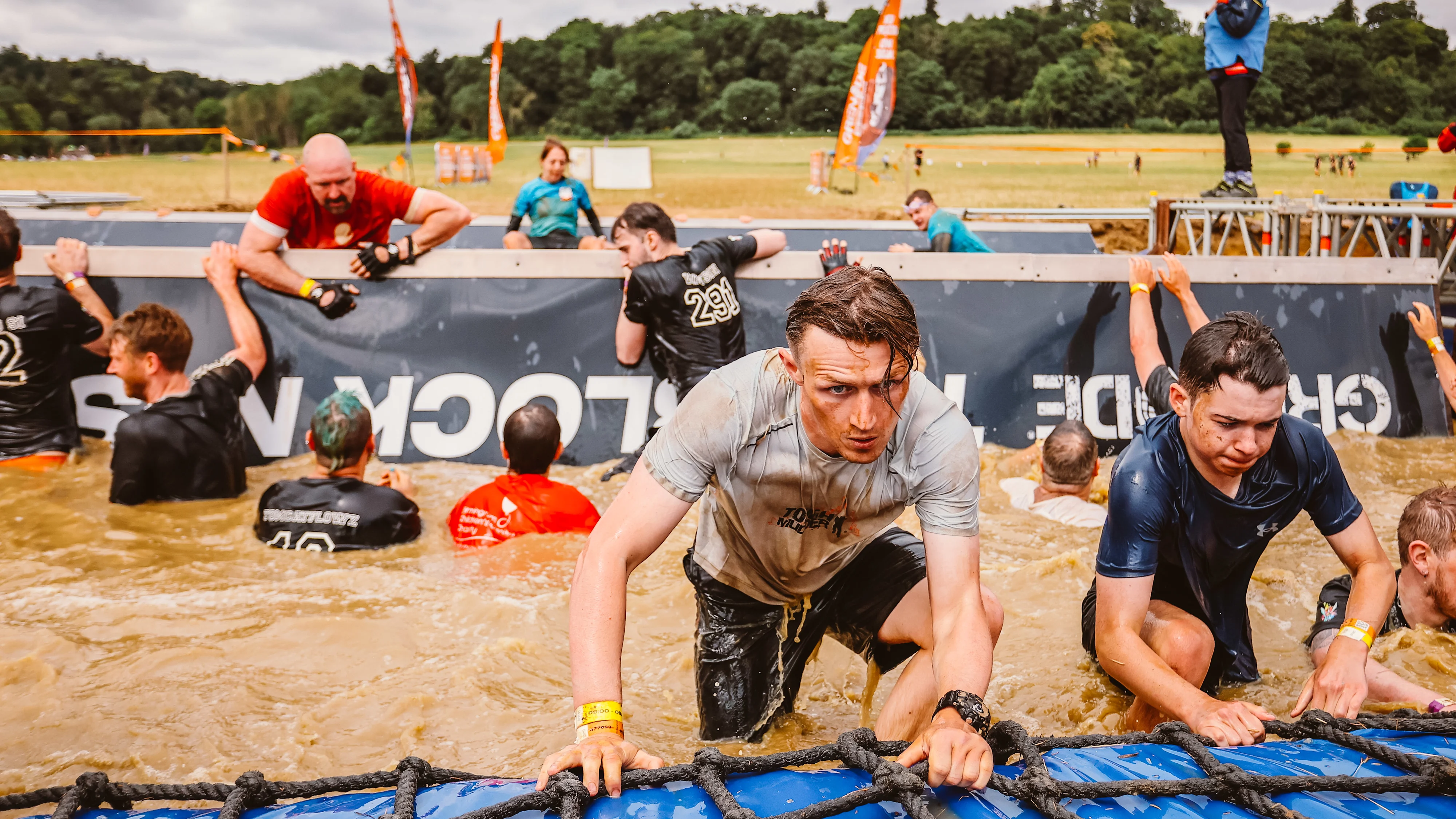 A Tough Mudder event outside in a field. A group of people are in a large pool of muddy water, some are climbing out of the sides holding onto ropes.